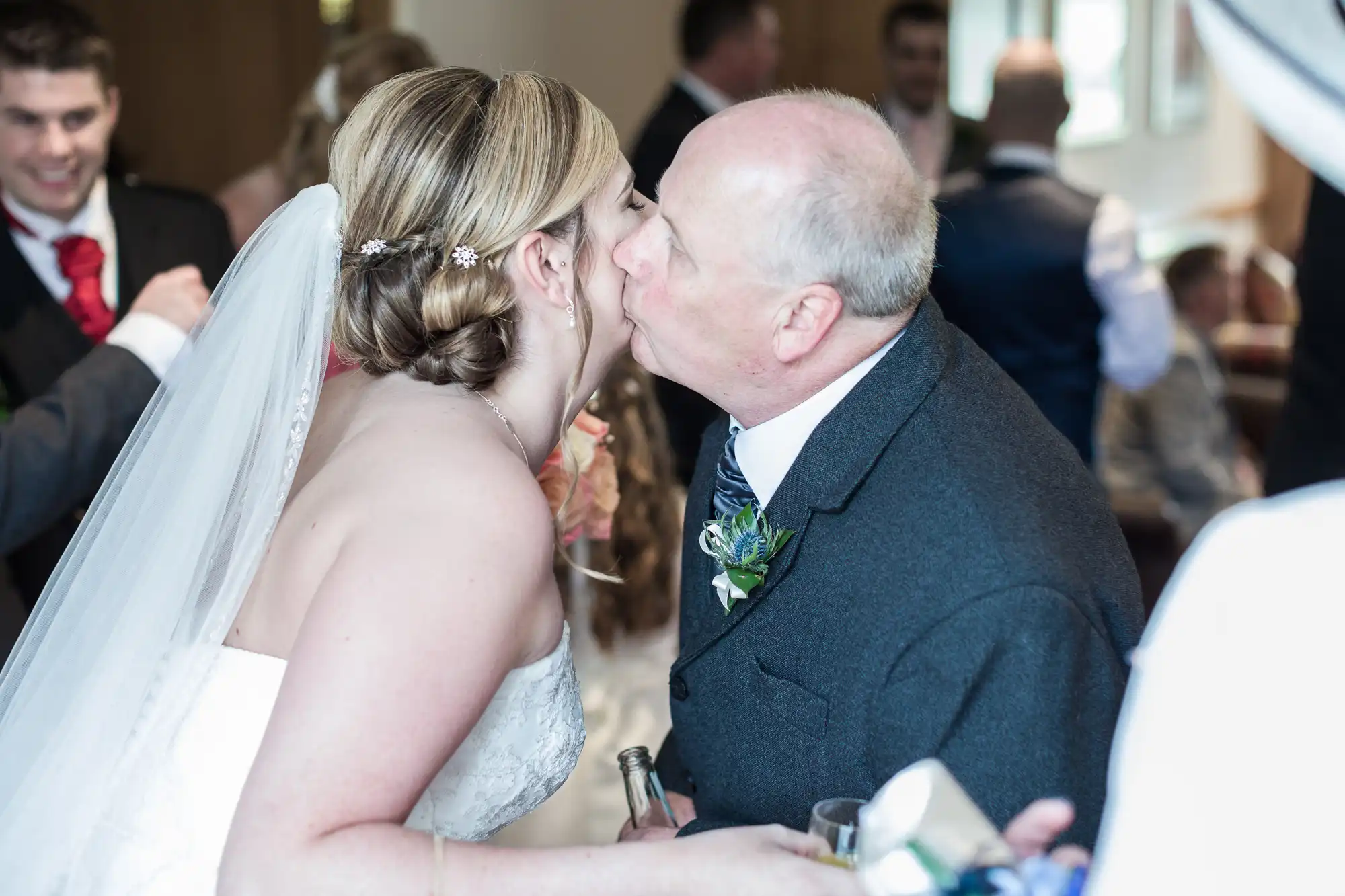 A bride in a white dress and veil receives a kiss on the cheek from an older man in a suit, likely her father. They are smiling warmly in a bright, crowded room with guests in the background. The mood is joyful and celebratory.