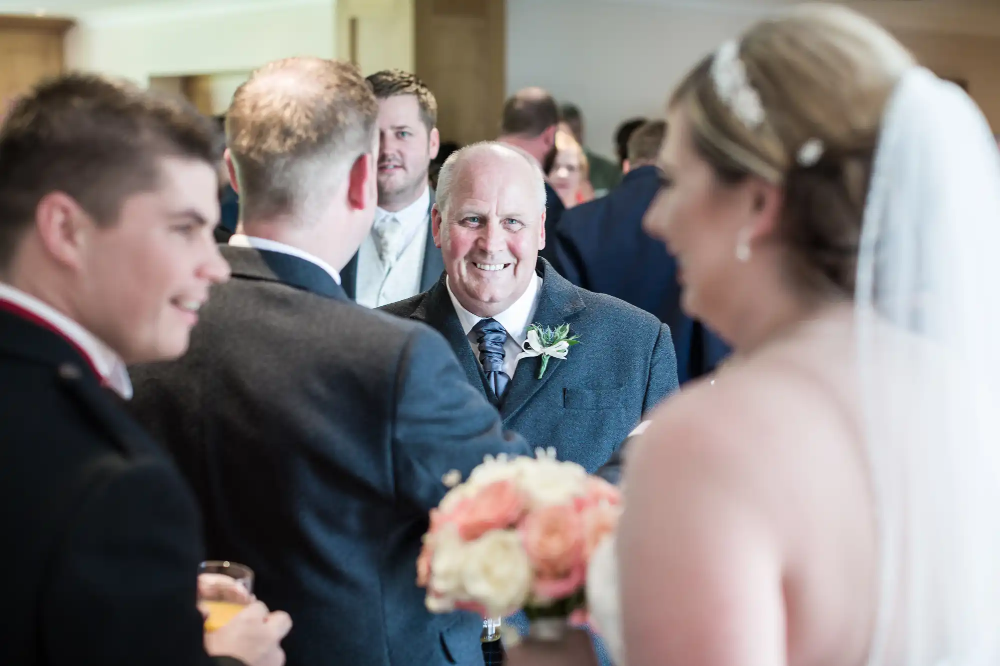 A smiling older man in a suit and buttonhole stands among a group at a wedding reception. Blurred in the foreground is a bride in a veil holding a bouquet, and other guests in formal attire are gathered, talking and holding drinks.