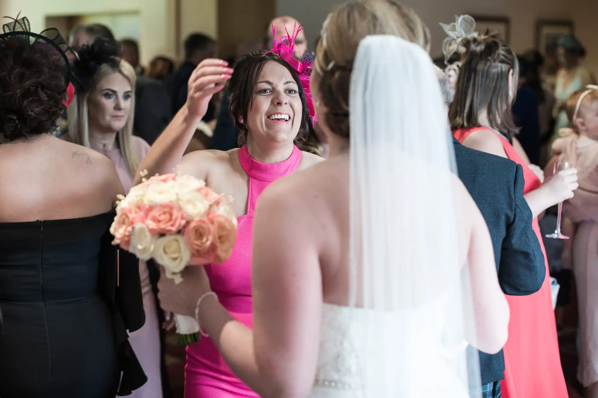 A bride in a white gown and veil holds a bouquet of roses while facing a smiling woman in a bright pink dress and fascinator, who raises her arms to greet her. Other guests in dresses and hats mingle in the softly lit background.