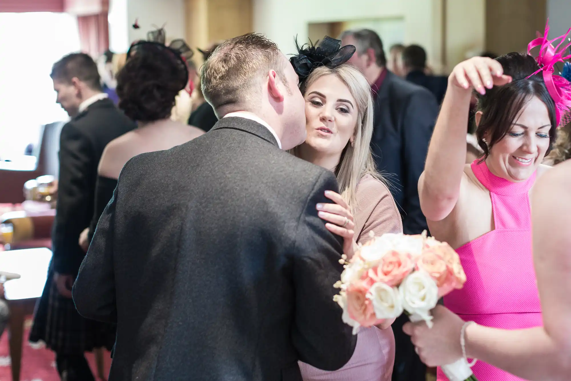 A man in a suit kisses a smiling woman on the cheek at a lively indoor event. She holds a bouquet of white and peach roses. Another woman in a bright pink dress and fascinator smiles, raising her arm. Other guests mingle in the softly lit background.