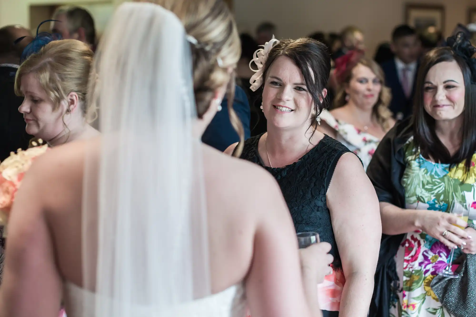 A bride in a white dress and veil faces a smiling woman in a black dress at a lively indoor wedding reception. Several other guests, both men and women, stand nearby, chatting and dressed in colourful formal attire.