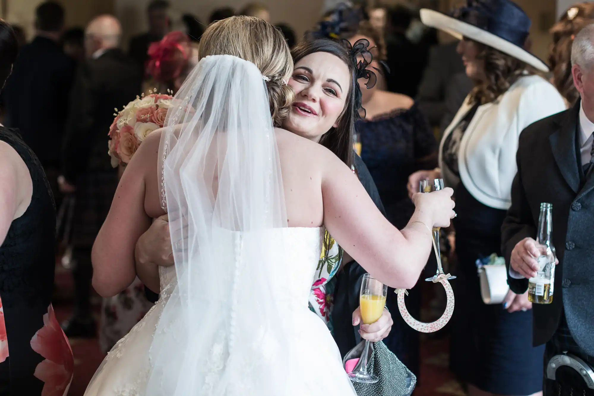A bride in a white dress and veil hugs a smiling woman at a wedding reception. The woman holds a drink and a handbag. Other guests in formal attire, some with hats, are visible in the background. The scene is celebratory and joyful.