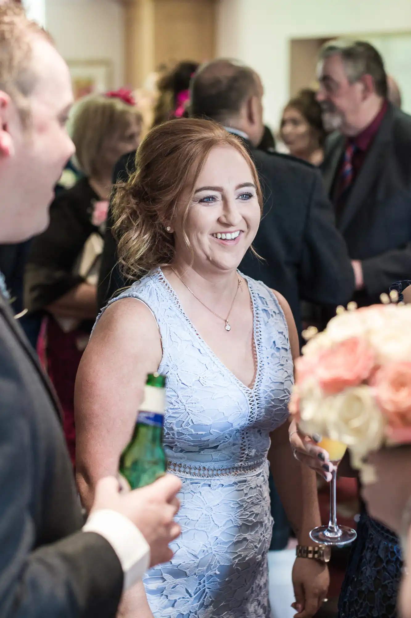 A woman in a light blue lace dress smiles whilst holding a drink at a social gathering. She is surrounded by other people, some holding drinks, and a bouquet of pink and cream flowers is visible in the foreground. The atmosphere is lively and cheerful.