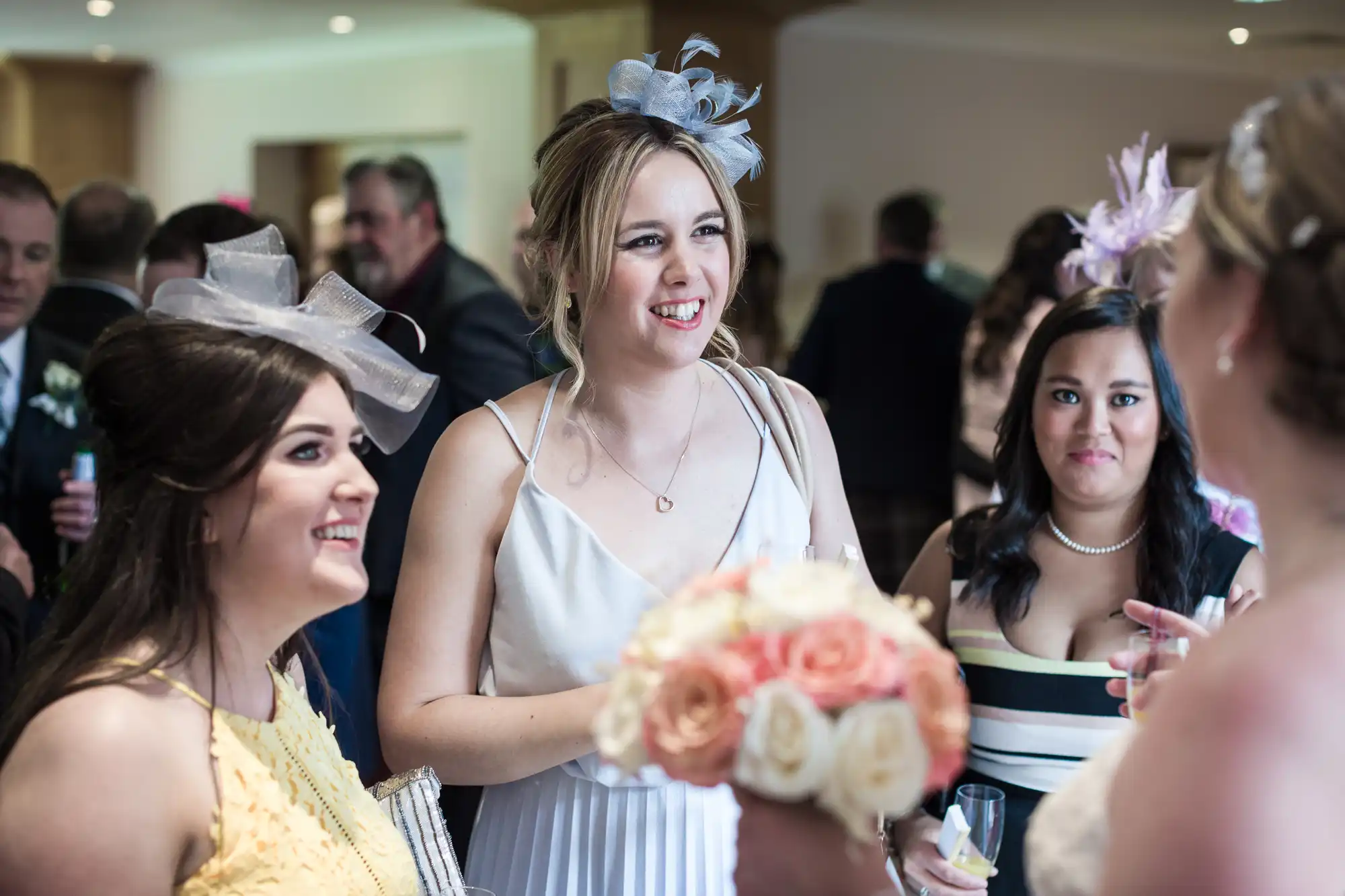 Three women in elegant dresses and fascinators smile and talk at a wedding reception. One holds a bouquet of roses. Other guests and soft natural light fill the blurred background, creating a joyful, celebratory atmosphere indoors.