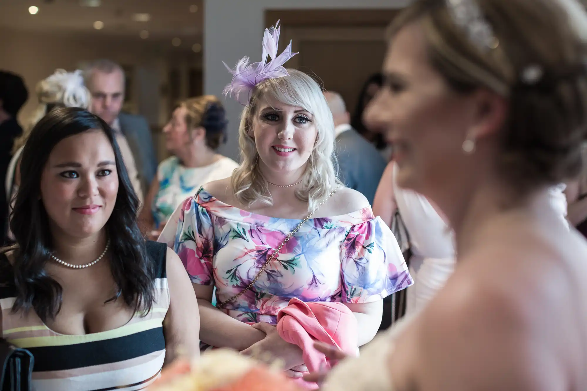 Two women, one in a striped dress and pearls, the other in a floral dress with a lavender fascinator, stand indoors among wedding guests, both looking towards a bride in the foreground, who is out of focus and holding a bouquet.