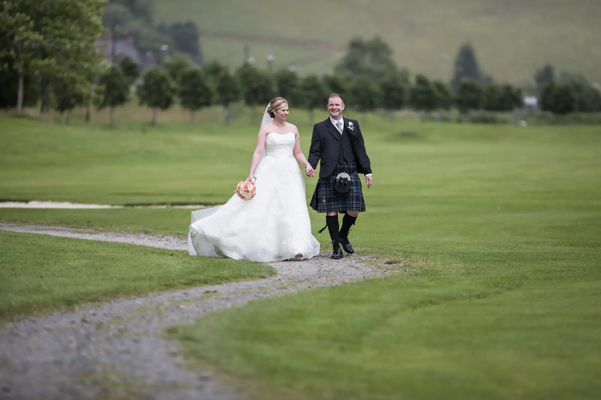 A bride in a white gown and a groom in a black jacket, waistcoat, and kilt walk hand in hand on a gravel path across a green lawn, smiling at each other. Trees and a hillside are visible in the blurred background.