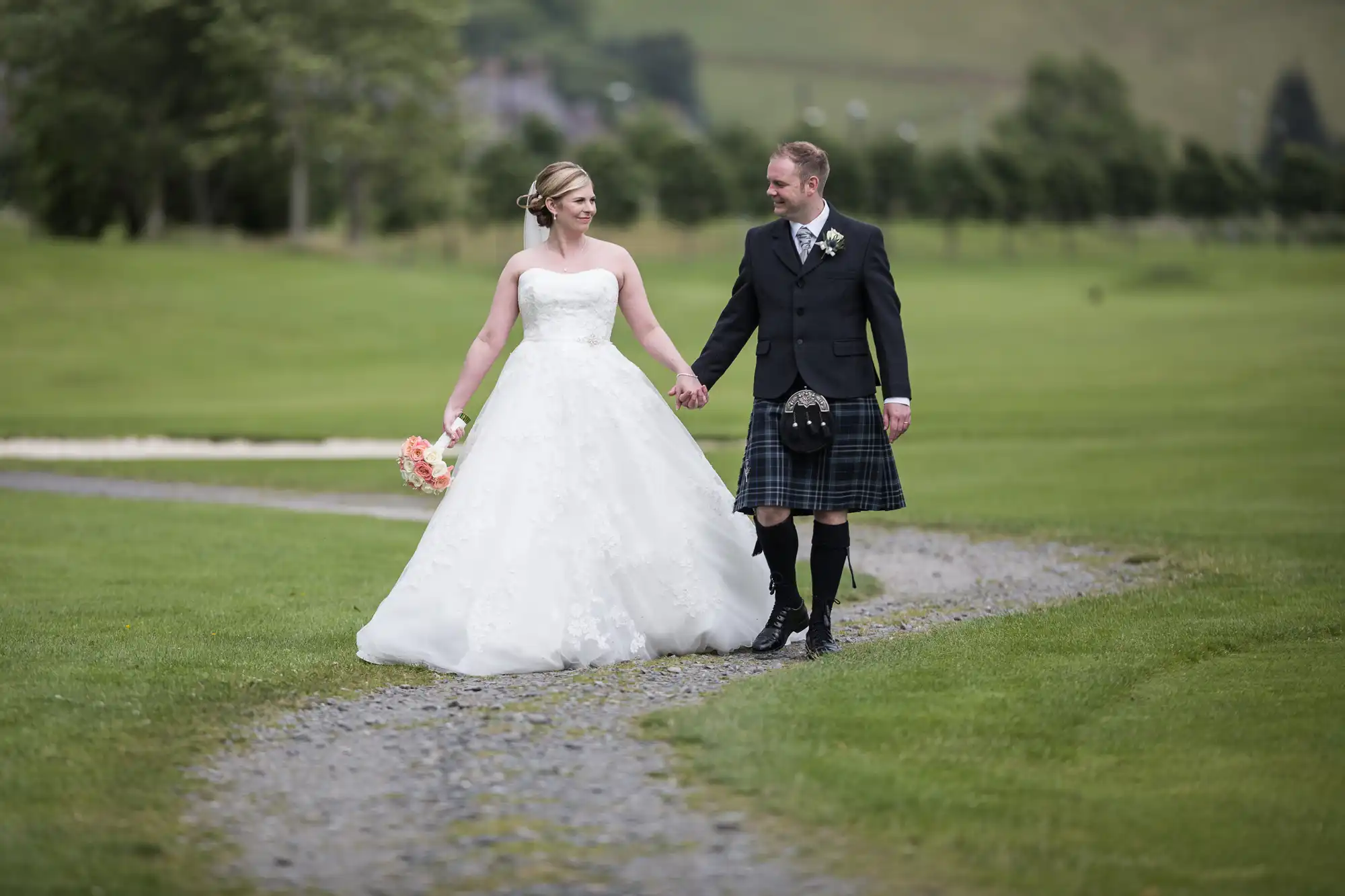 A bride in a white dress holding a bouquet and a groom in a black jacket and tartan kilt walk hand-in-hand along a gravel path on a green lawn, smiling at each other, with trees and hills in the background.