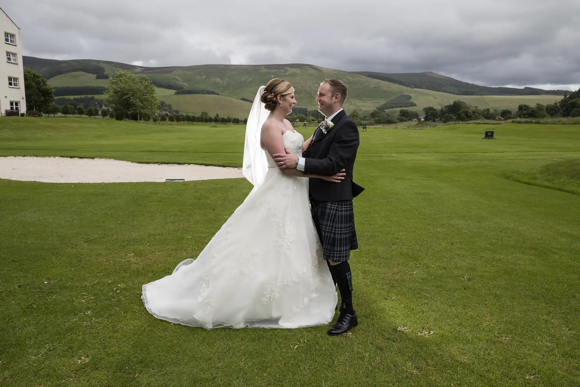 A bride in a white gown and veil embraces a groom in a black jacket and tartan kilt on a green lawn, with rolling hills, trees, and cloudy sky in the background. They look at each other, smiling, near a sand bunker on a golf course.