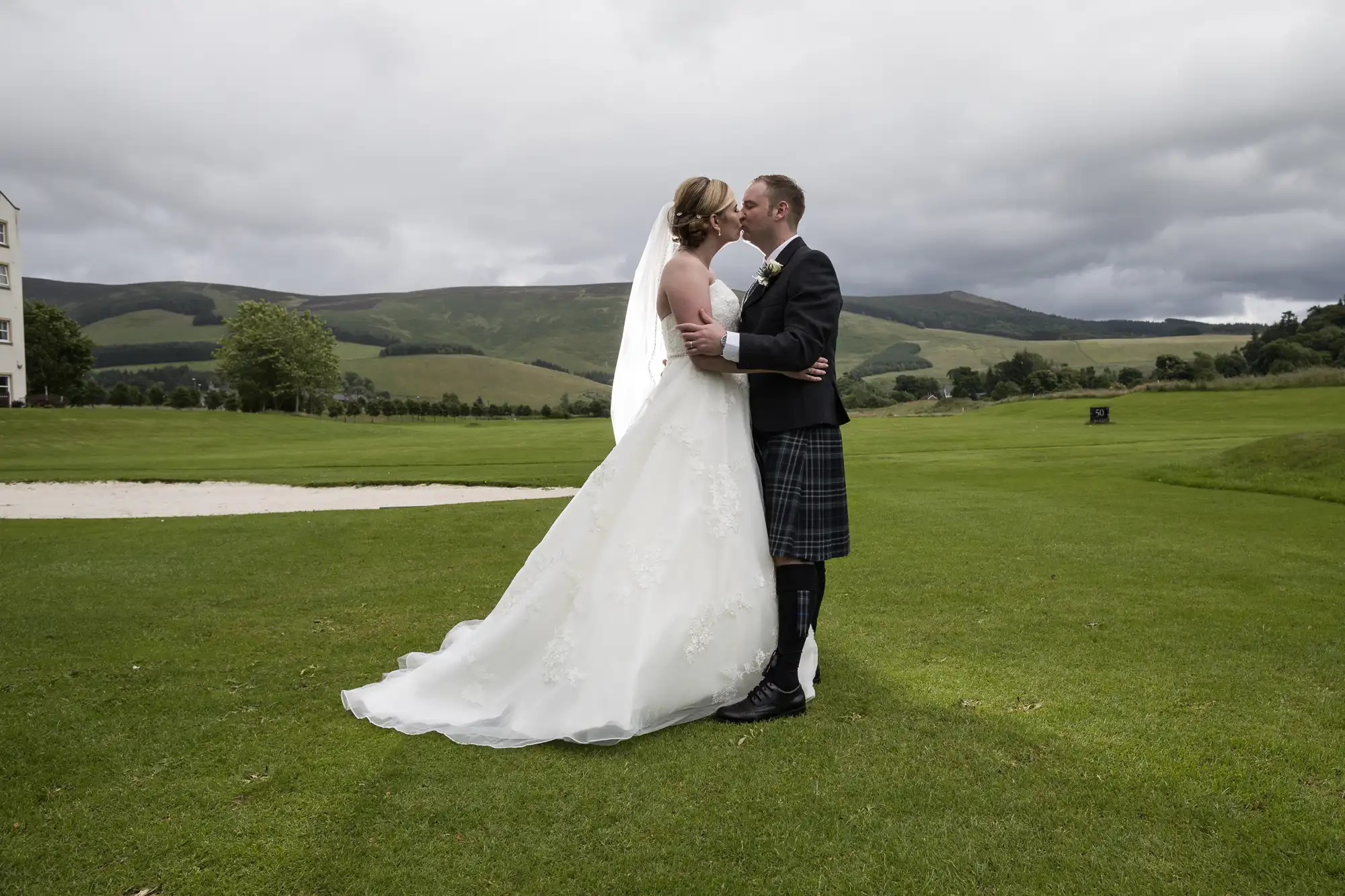 A bride in a white gown and veil embraces a groom in a traditional Scottish kilt on a grassy golf course, with rolling hills and cloudy skies in the background. They stand close, facing each other, appearing about to kiss.