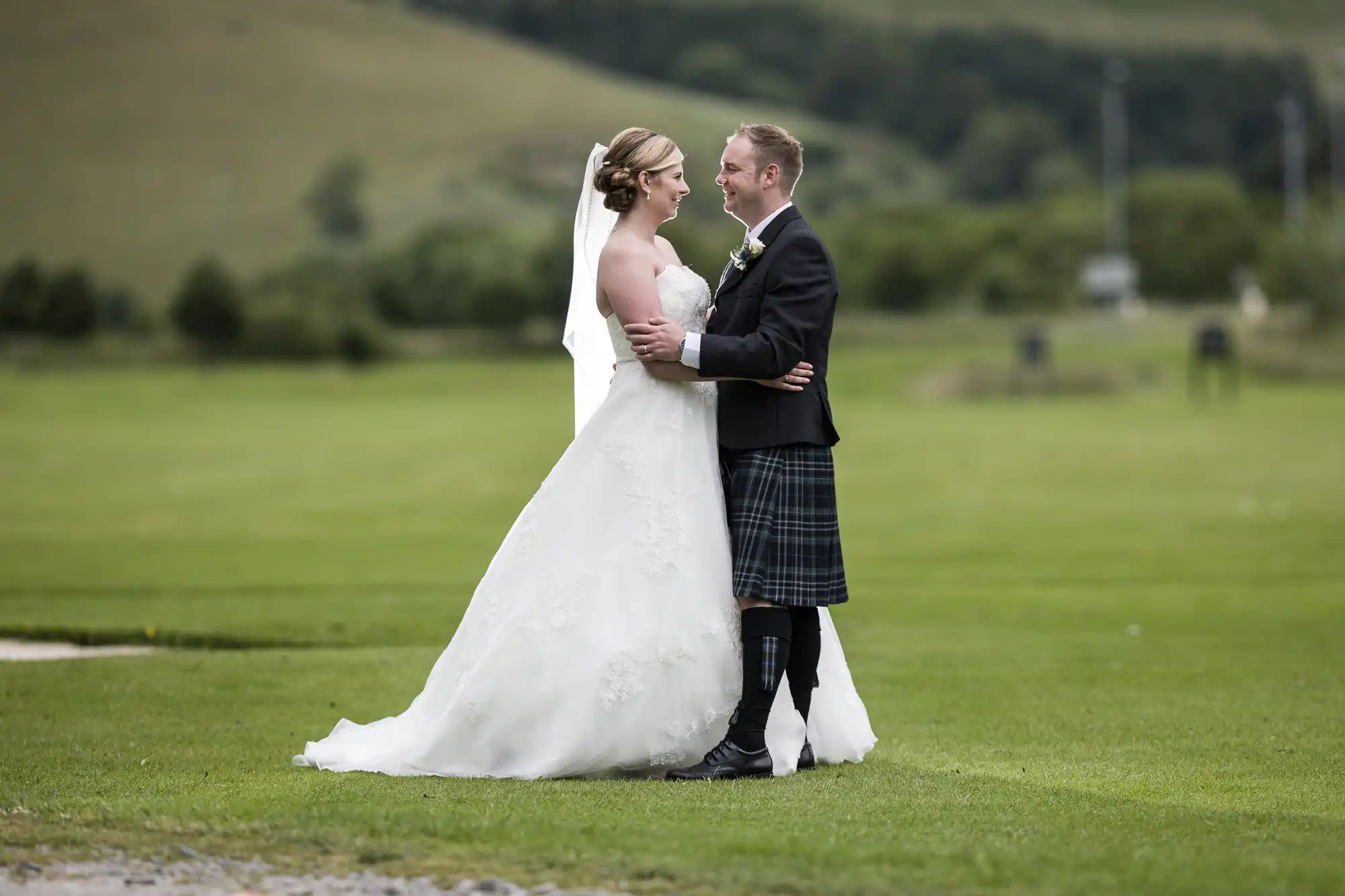 Macdonald Cardrona Hotel wedding photographer, Love Wedding Photos And Film colour image of a bride in a white gown and veil embraces a groom in a black jacket, kilt, and knee-high socks. They stand on green grass with rolling hills and trees in the background, smiling at each other in an outdoor setting.