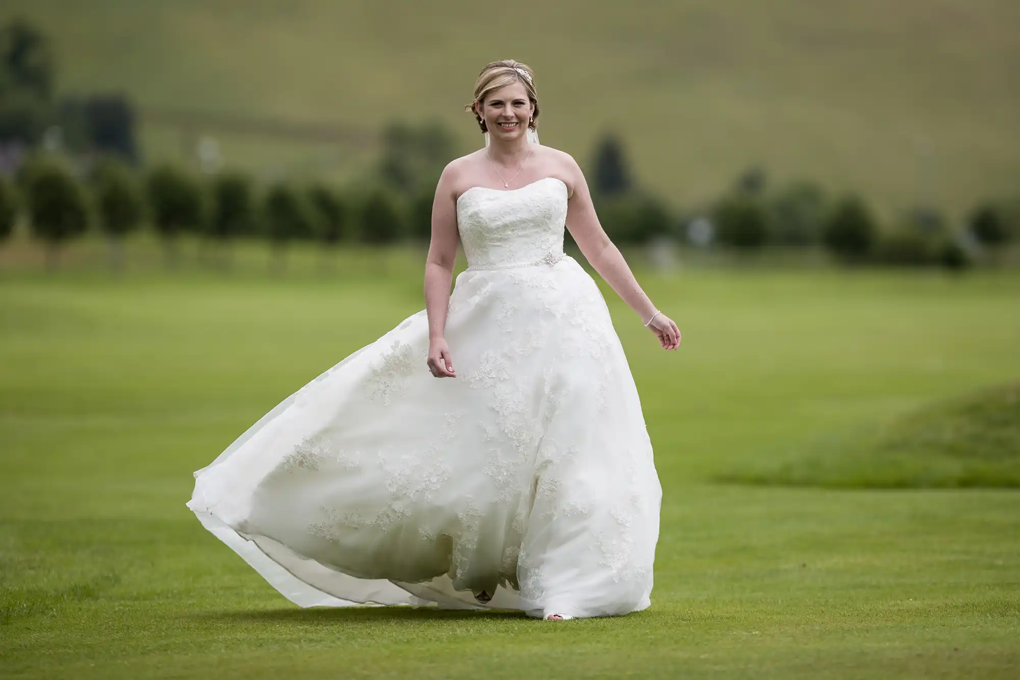 A woman in a strapless white wedding dress stands on a green lawn, smiling. She has fair hair styled up and wears a tiara. The dress has a full, lacy skirt that flows outward. The blurred background shows distant trees and grassy hills.
