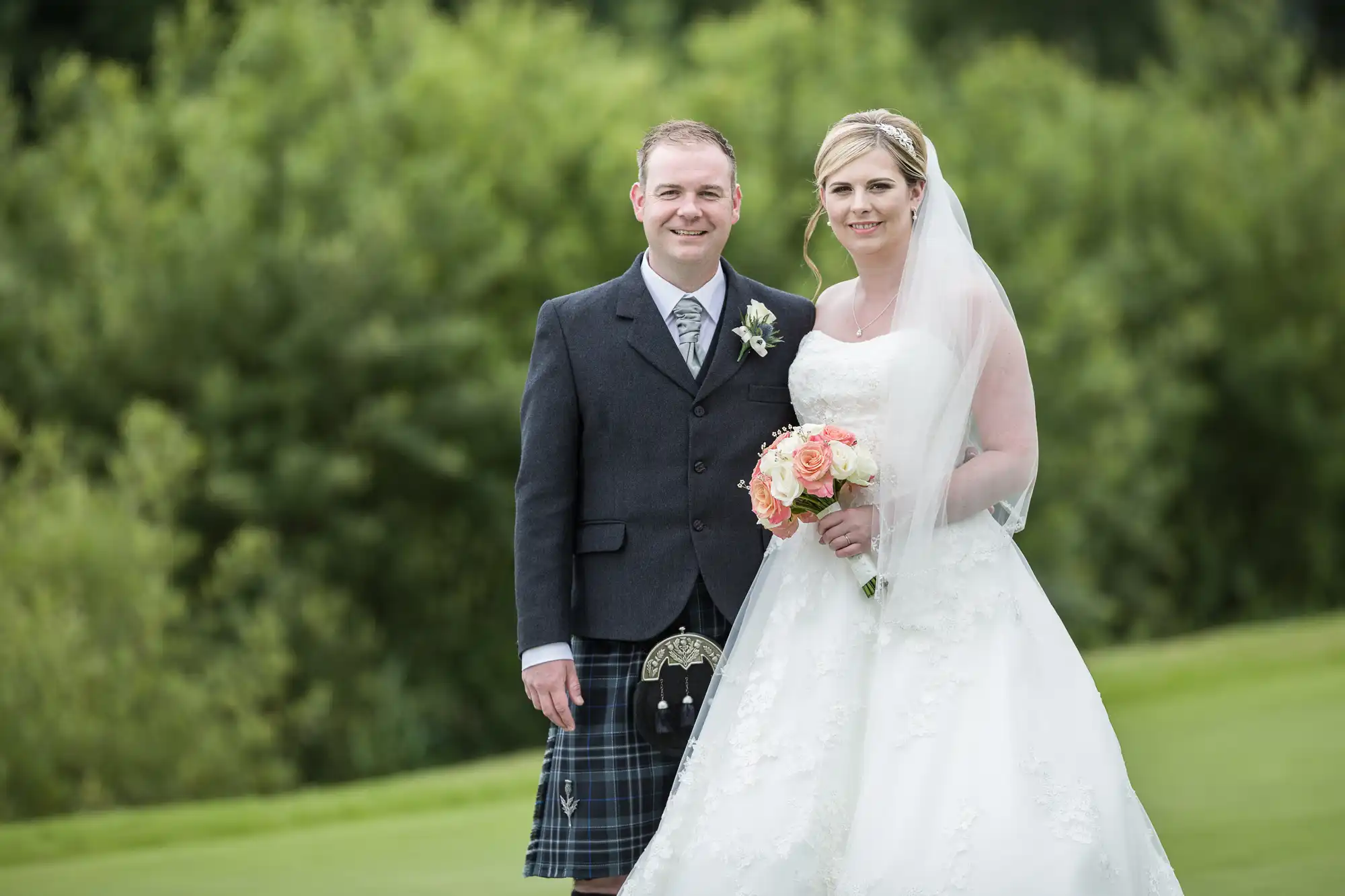 A smiling bride in a white dress and veil holds a bouquet of pink and white flowers, standing beside a groom in a dark jacket and traditional Scottish kilt. They pose outdoors on grass with green, leafy trees in the background.