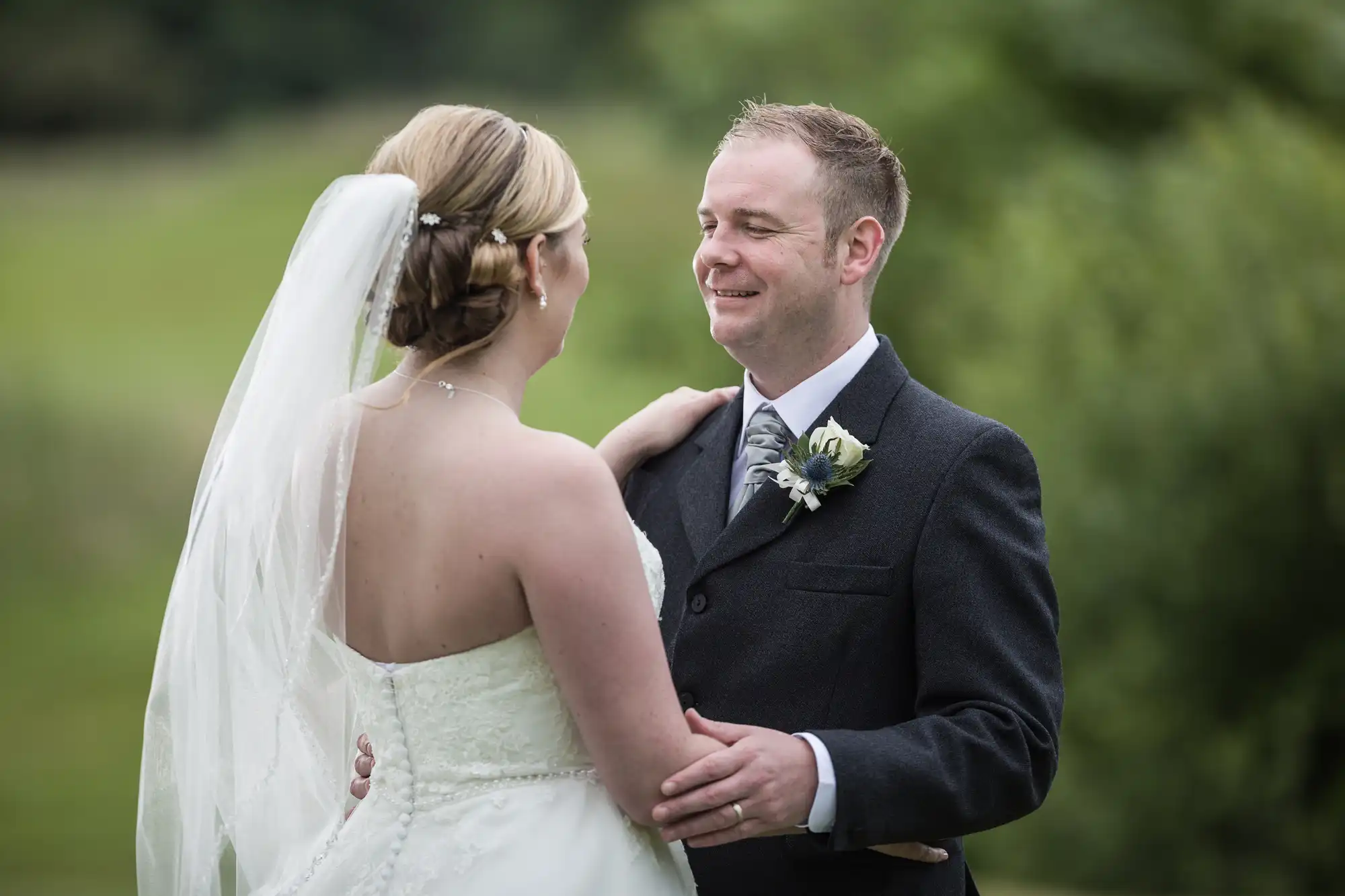 A bride in a strapless white dress and veil embraces a groom in a dark suit with a buttonhole. They smile at each other outdoors, surrounded by greenery and blurred natural scenery.