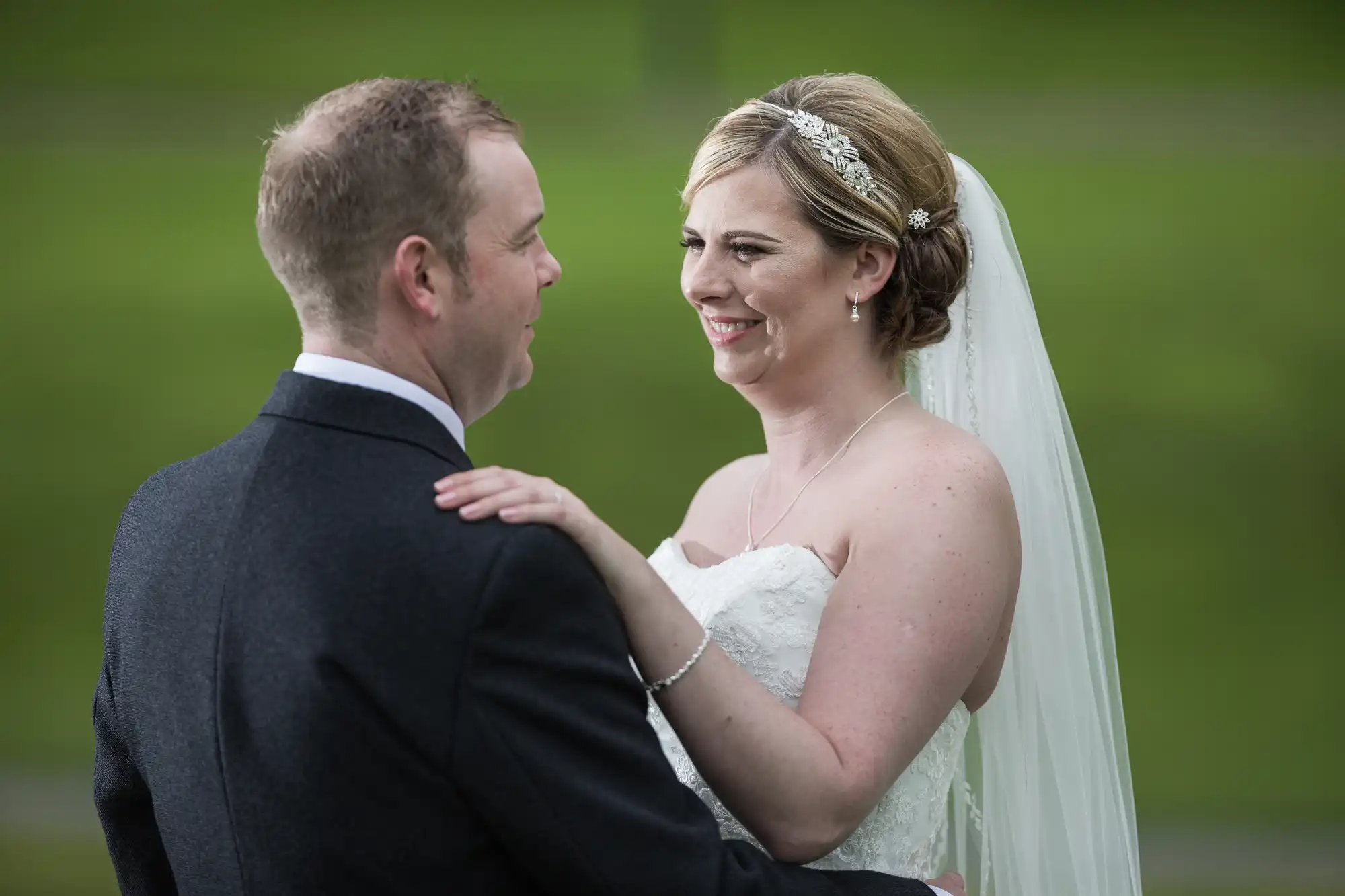 A bride in a white strapless dress and veil smiles at the groom in a dark suit as they embrace outdoors. The bride wears a jewelled headband and necklace; both look happy with green grass blurred in the background.