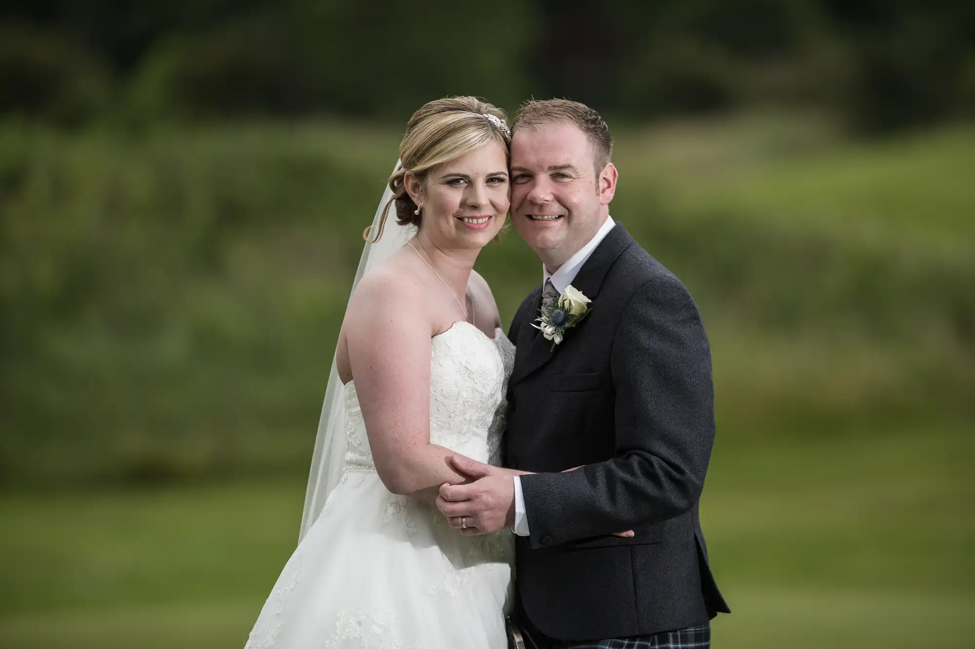 A bride in a white strapless gown and veil stands close to a groom in a dark jacket and checked trousers. Both are smiling, arms round each other, with a blurred green outdoor background behind them.