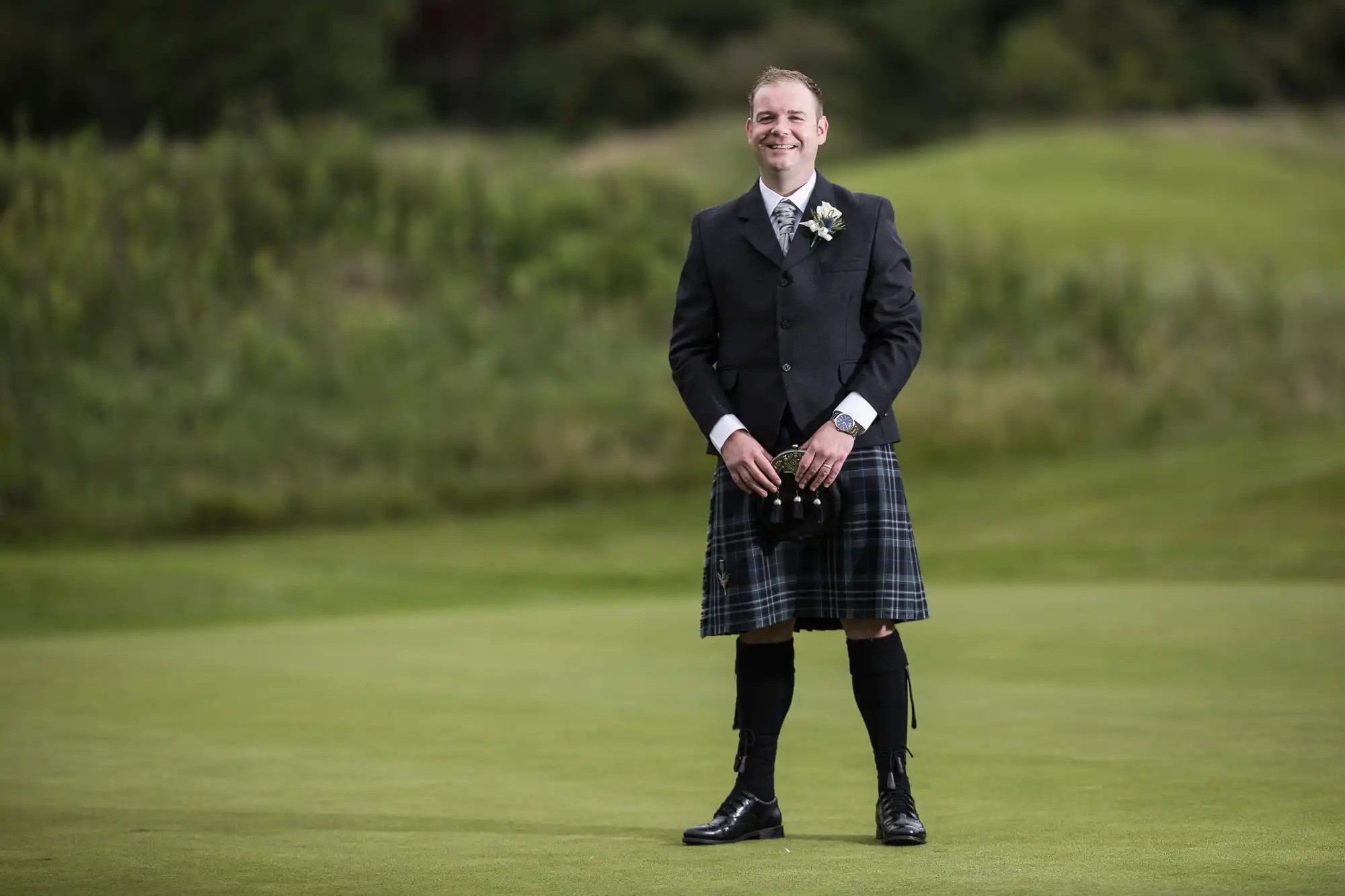 A smiling man stands on grass, wearing a dark jacket, white shirt, tartan kilt, knee-high socks with flashes, and black shoes. He holds a sporran and has a buttonhole on his lapel. The background shows blurred green trees and fields.