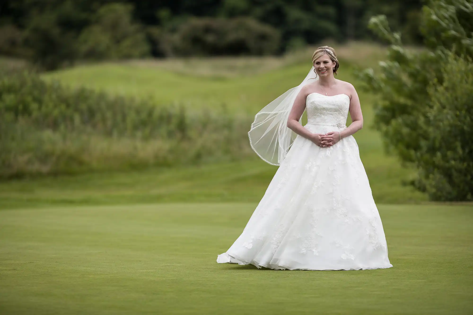 A bride in a strapless white wedding dress with lace details and a veil stands smiling on a green lawn, hands clasped, with a blurred background of trees and shrubs on a cloudy day.