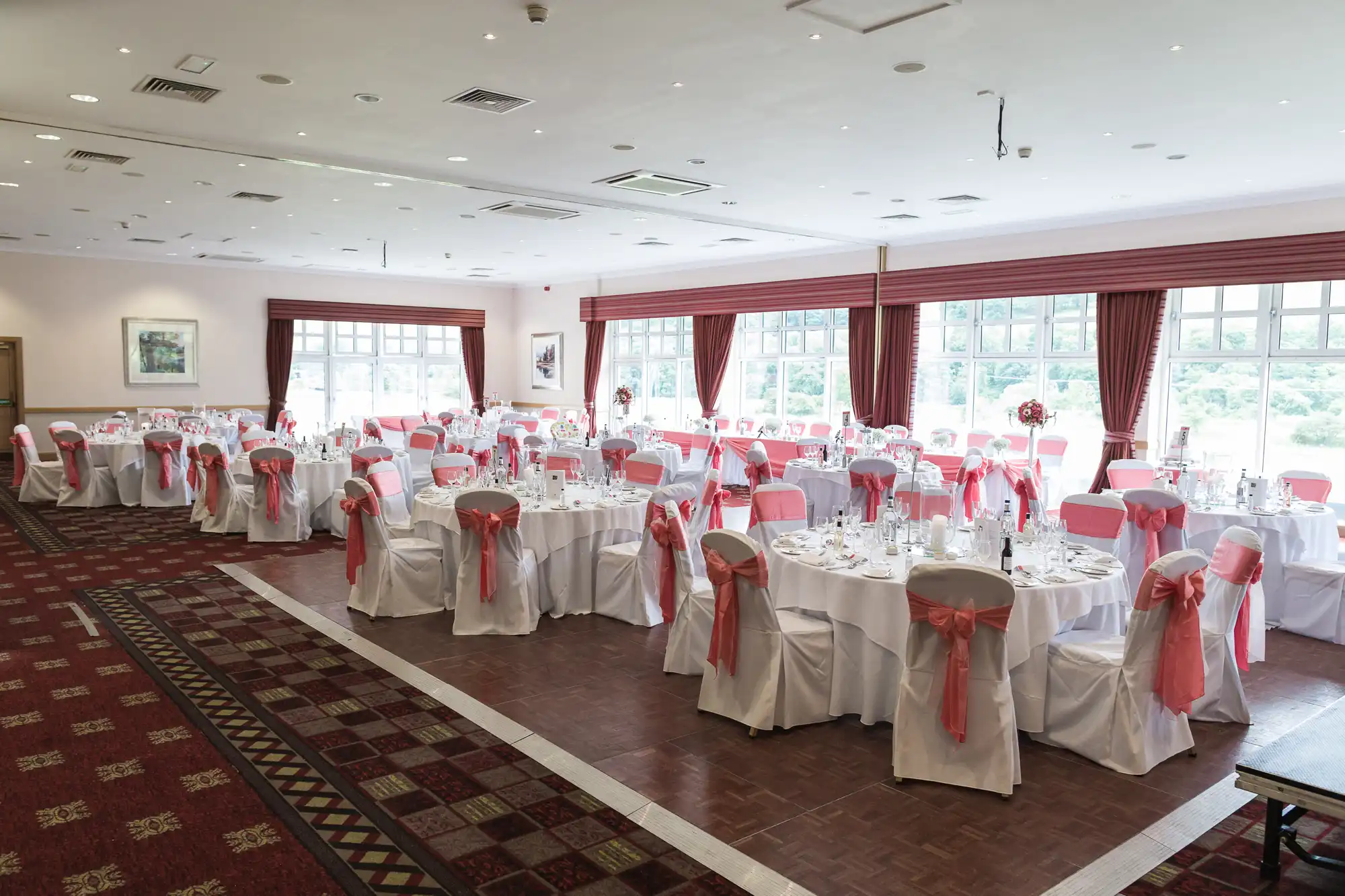 A spacious banqueting hall is set for an event, with round tables covered in white cloths and white chairs adorned with pink sashes. Large windows let in natural light, and red curtains frame the windows. Place settings and glassware are neatly arranged on each table.