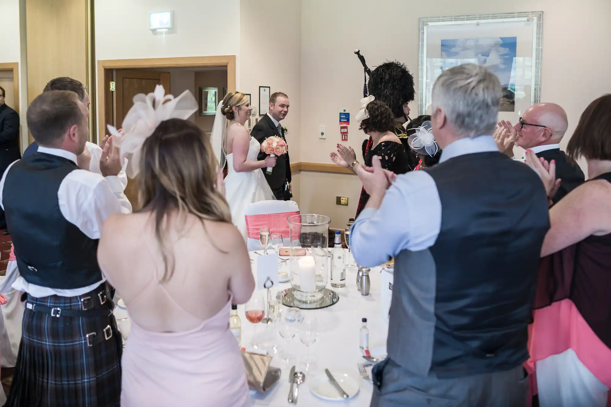 A bride and groom enter a wedding reception as guests stand, clap, and smile. The bride holds a bouquet, and the tables are set with glasses, bottles, and candles. People are dressed formally, with some women wearing decorative hats. The atmosphere is celebratory.