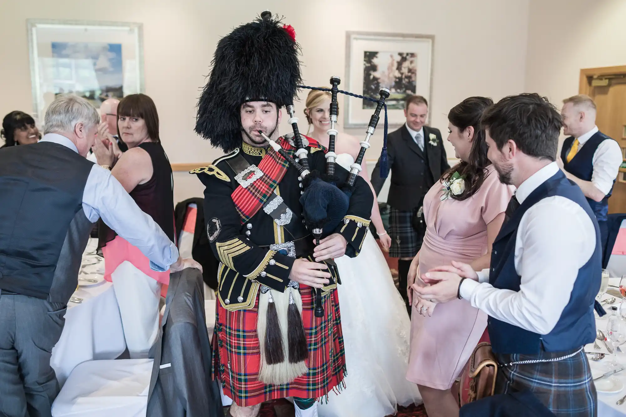 A bagpiper in traditional Scottish attire plays at a wedding reception. Guests in formal clothes, including kilts and dresses, gather around decorated tables. The bride in a white gown and groom are visible in the background, smiling and interacting with guests.