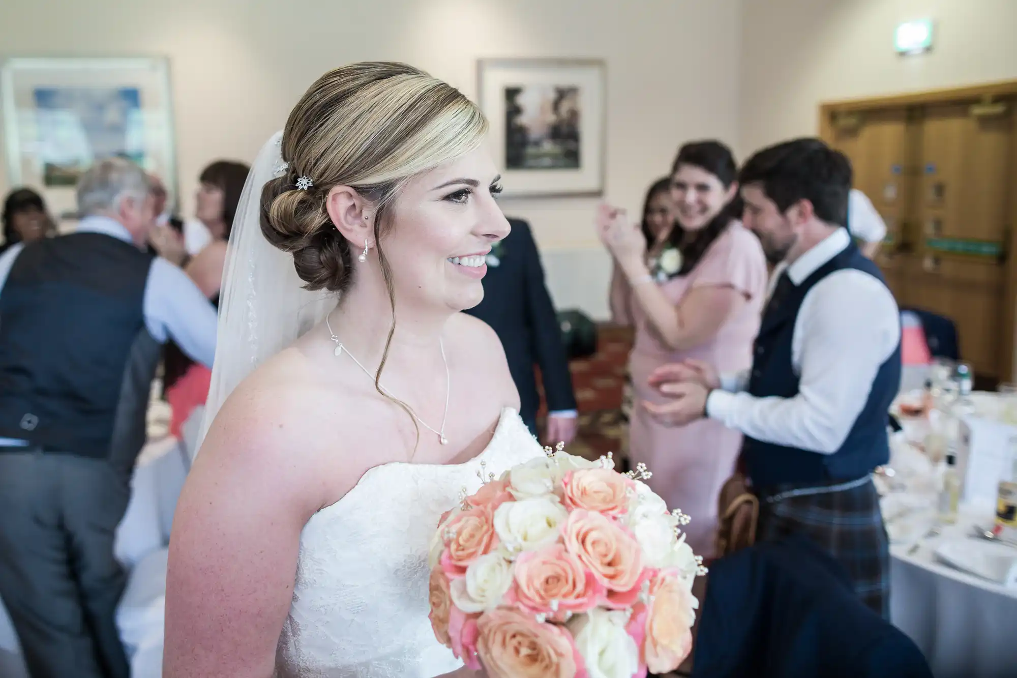 A smiling bride in a white dress and veil holds a bouquet of peach and cream roses. She stands indoors among guests dressed in formal attire, some applauding and chatting in a warmly-lit reception room.