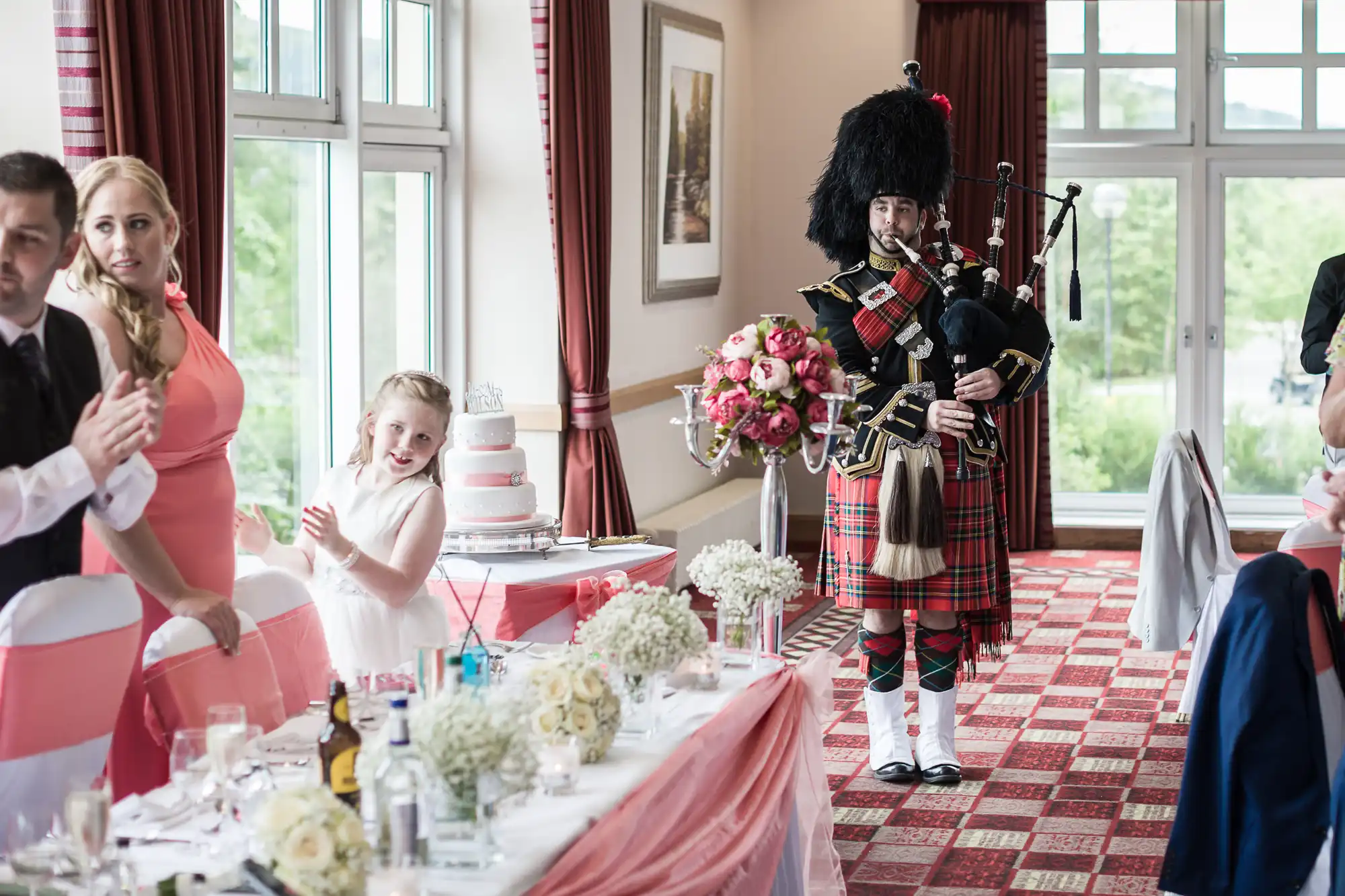 A man in traditional Scottish attire plays bagpipes by a table adorned with flowers and a pink cake. A young girl in white claps and smiles nearby, whilst guests in formal wear watch. Large windows and red curtains brighten the festive room.
