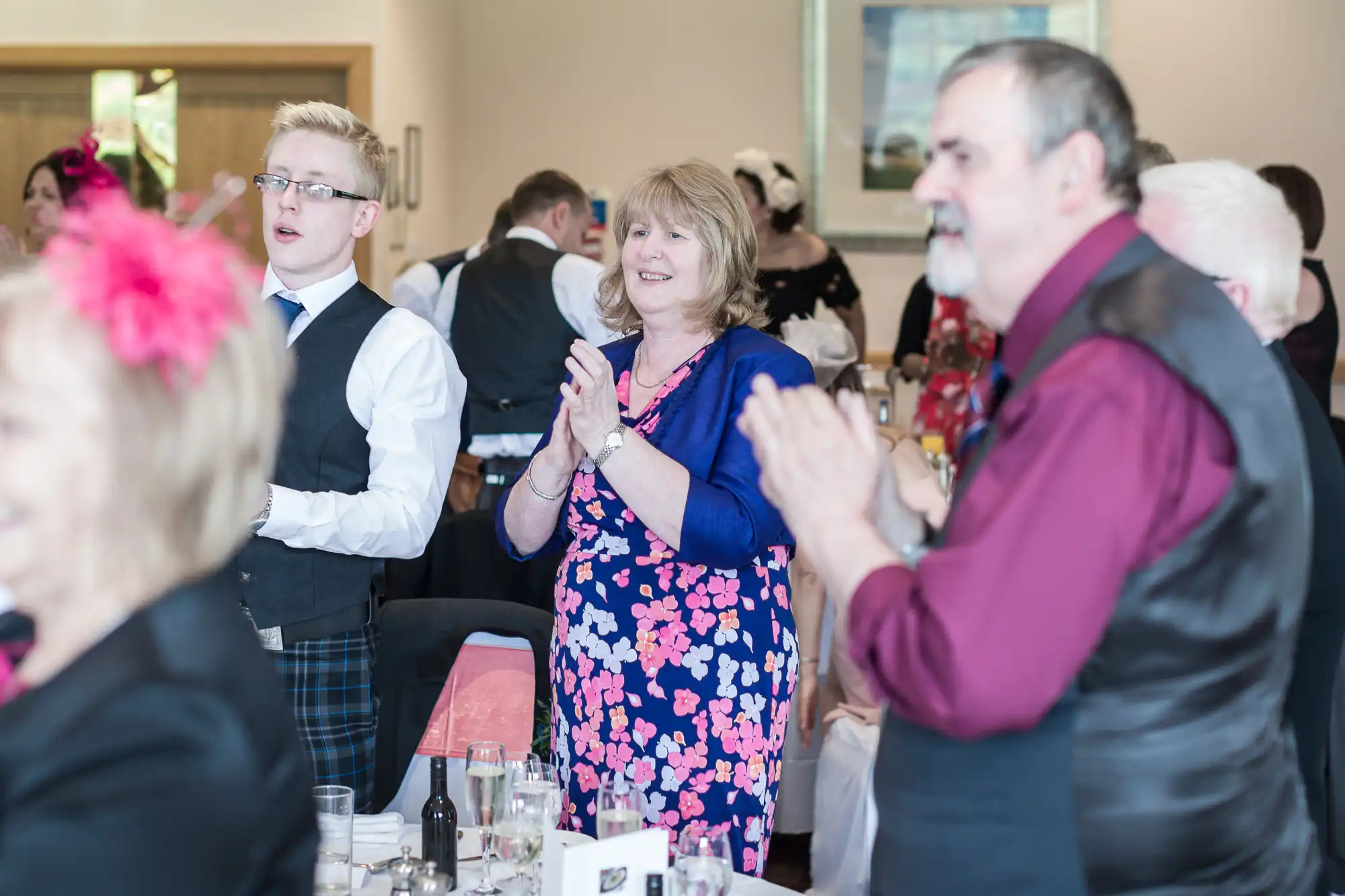 A group of people dressed formally, some standing and applauding at tables set for a celebration. A woman in a blue cardigan and floral dress is smiling and applauding in the centre. The atmosphere appears joyful and festive.