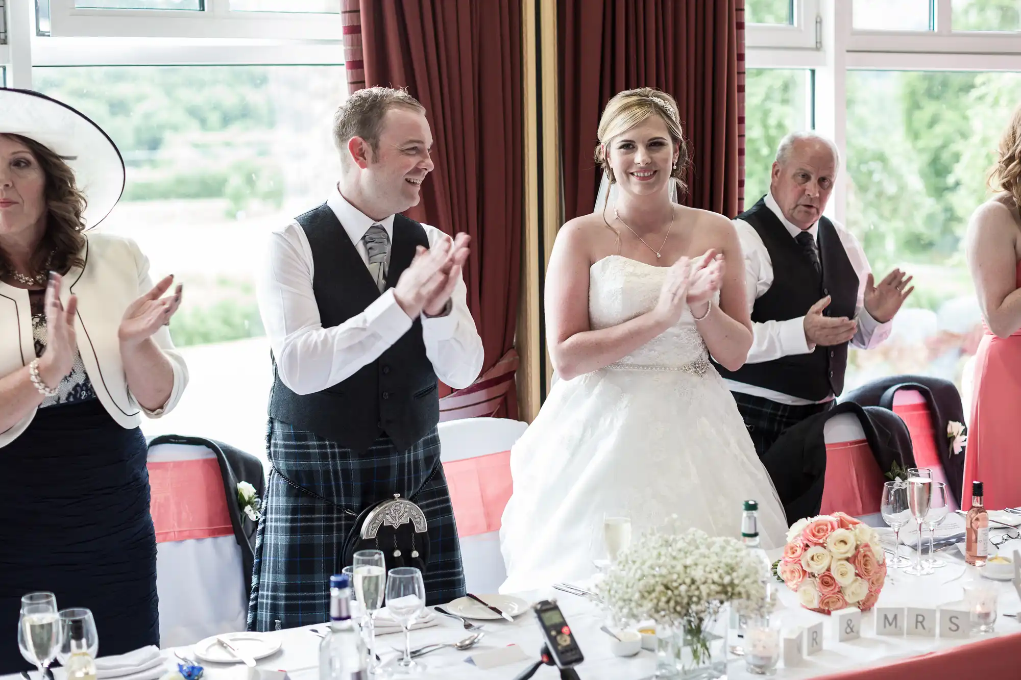 A bride in a white gown and a groom in a kilt stand clapping at a decorated wedding reception table, joined by guests in formal attire. The table has flowers, drinks, and a “MR & MRS” sign. Large windows and red curtains are in the background.