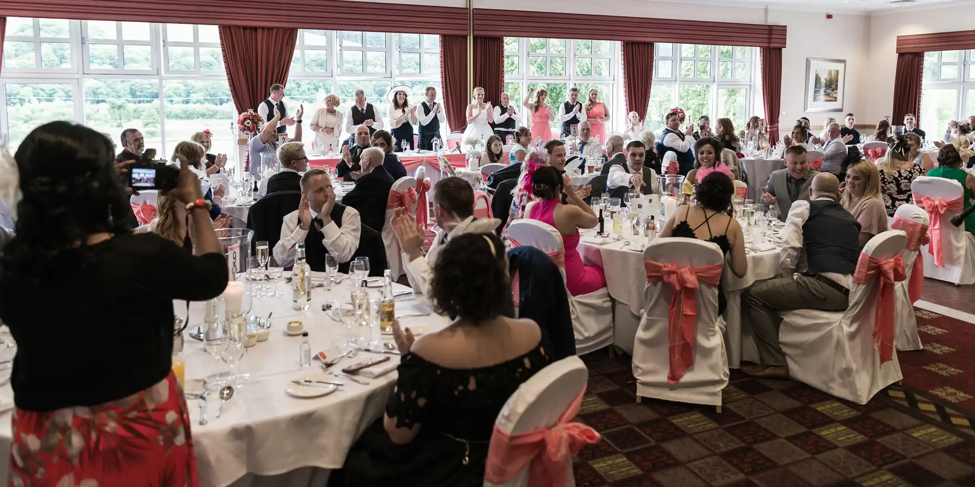 A large group of people sit at round tables covered with white cloths and pink chair sashes in a bright, windowed banqueting hall. Many are dressed formally. Some guests applaud, while others take photographs of a group of people standing at the far end of the room.