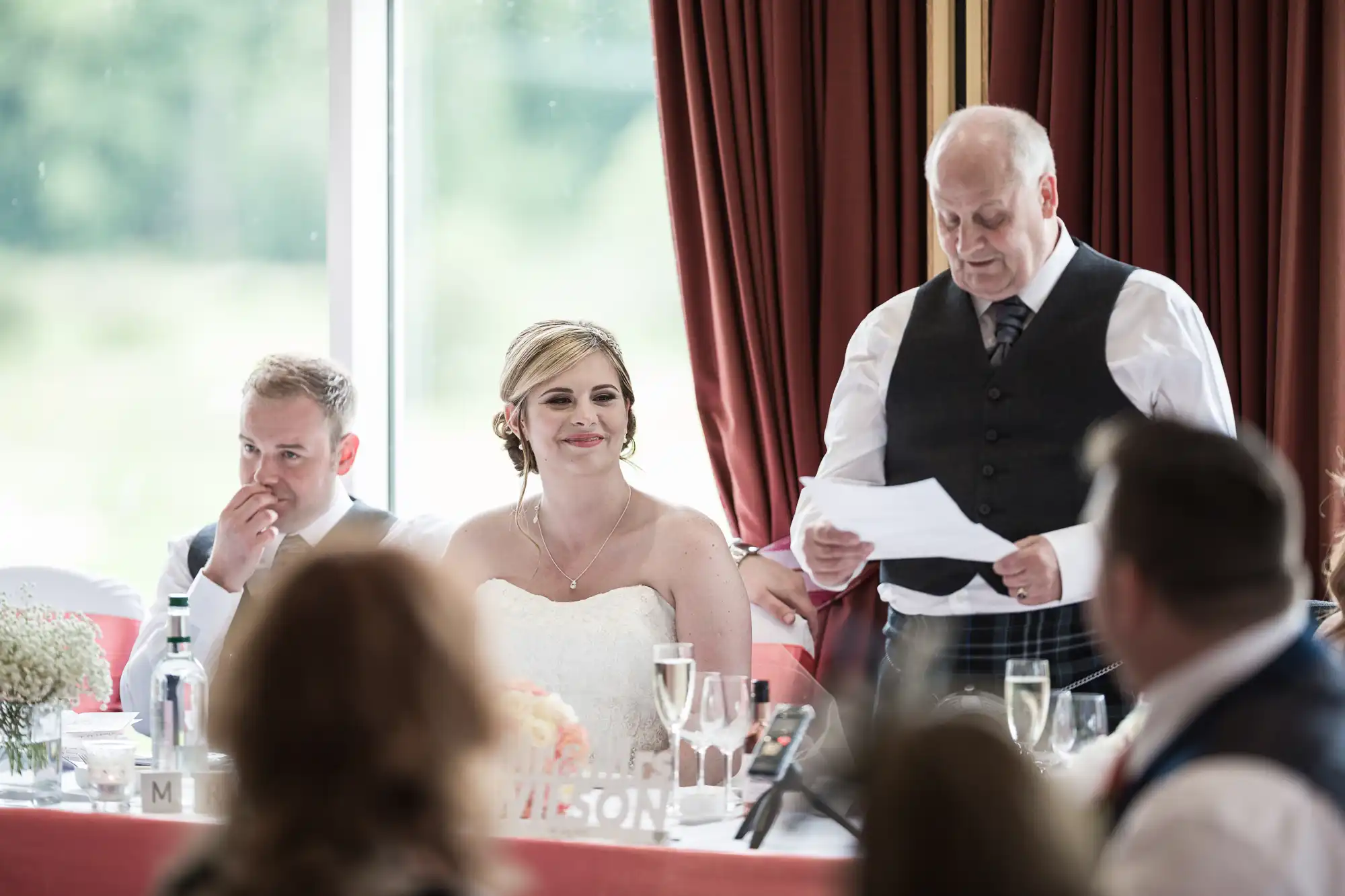 A bride in a white dress smiles as an older man in a waistcoat and tie reads from a piece of paper at a wedding reception. Guests sit at a long table decorated with flowers and glasses, with burgundy curtains and a window in the background.