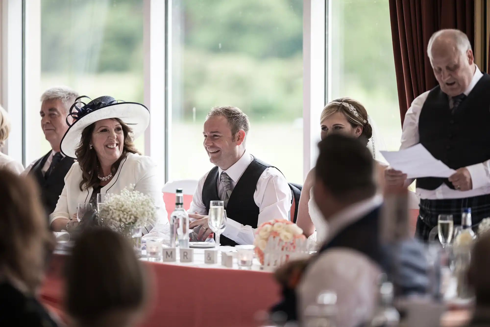 A wedding reception scene shows guests seated at a long table by large windows. A man in a waistcoat stands, reading from a paper, while others, including a woman in a large white hat and a smiling bridegroom, listen and smile. Champagne glasses and decorations are on the table.