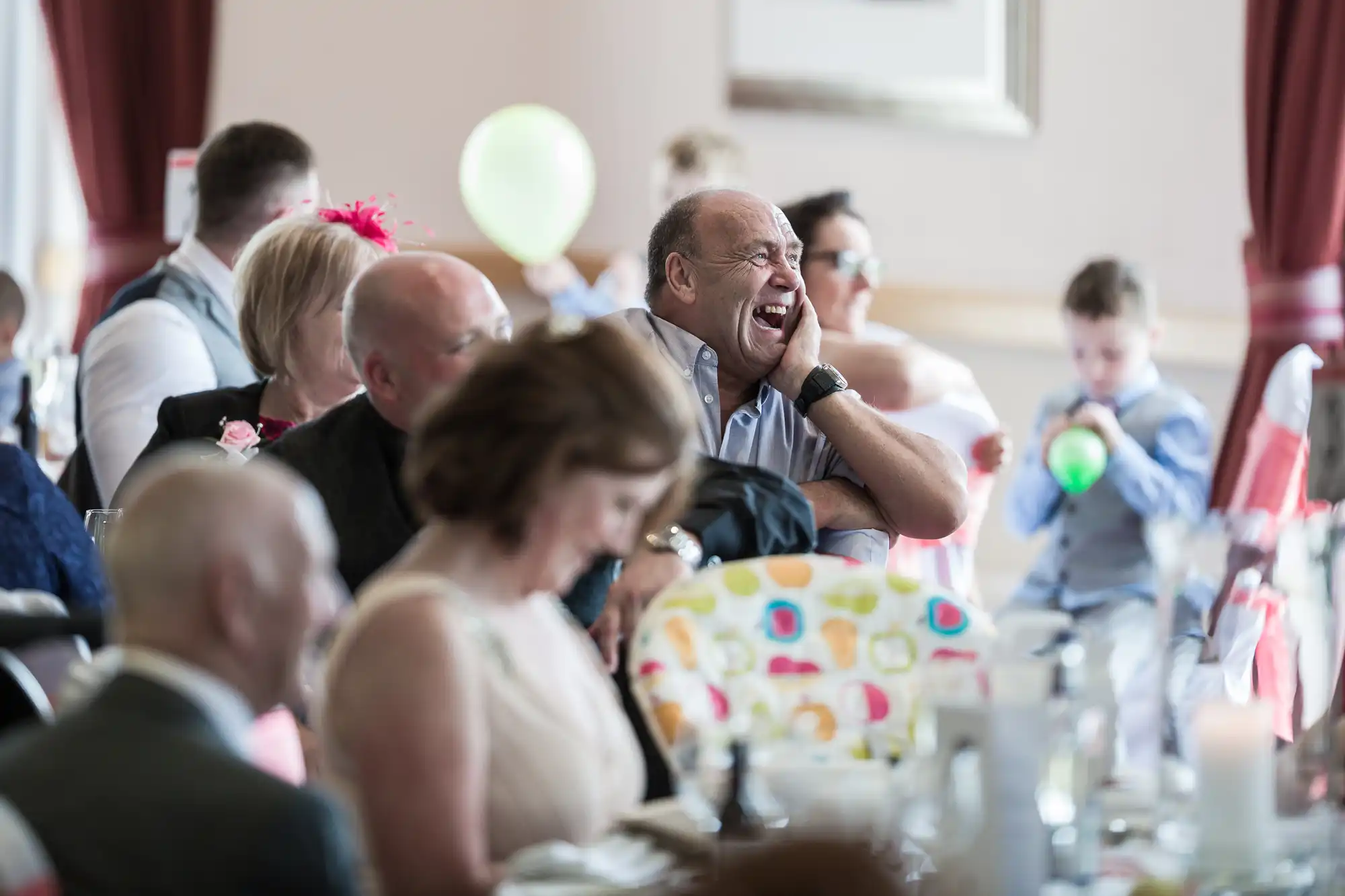 A man at a crowded banquet table laughs heartily, resting his head on his hand. Other guests, including children and adults, sit and interact nearby. The atmosphere appears festive and light-hearted, with balloons and decorations visible.