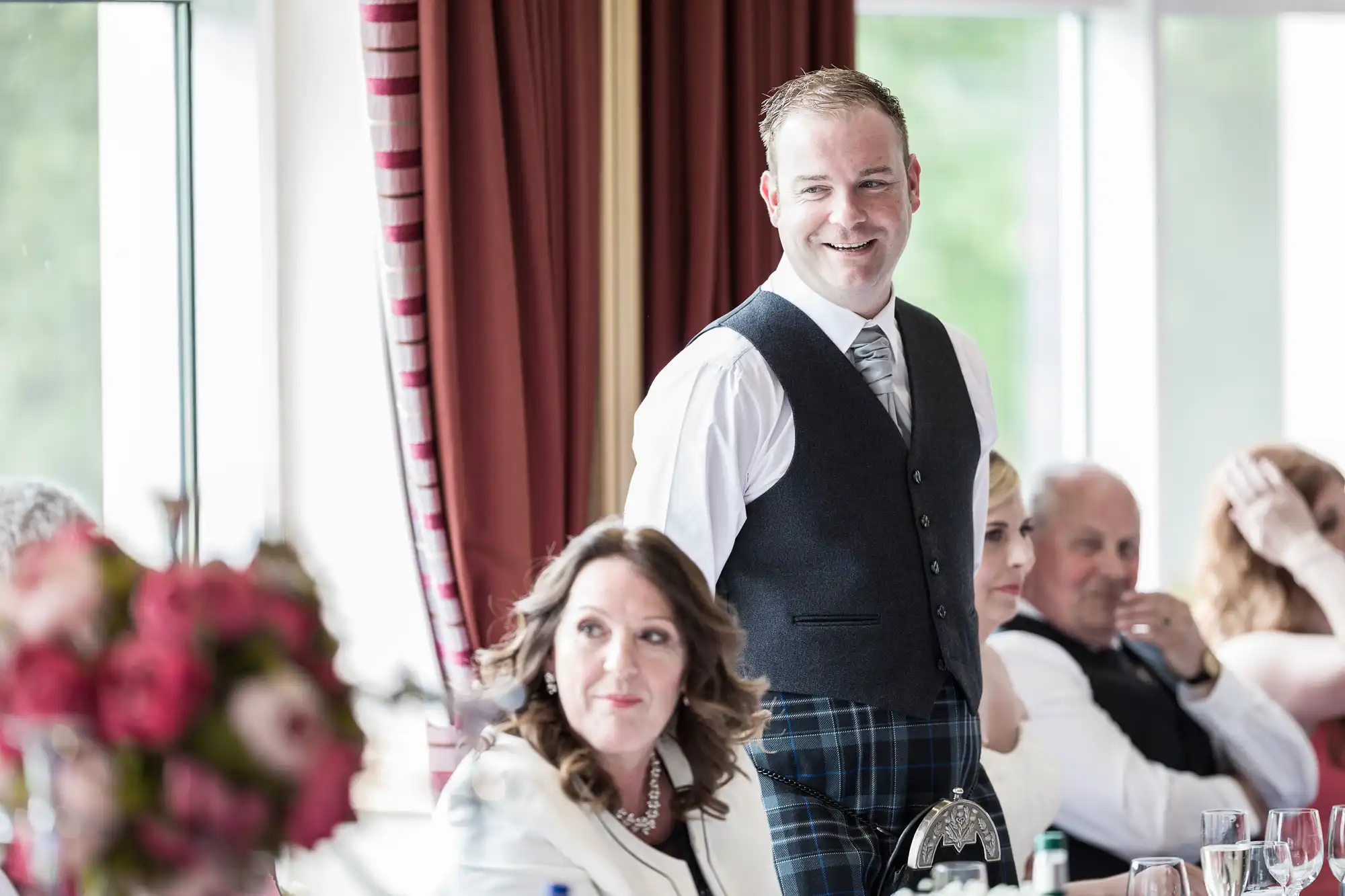 A man in a waistcoat and tie stands smiling at a formal event, possibly a wedding. Seated guests in formal attire listen, with a woman in focus in the foreground. Red curtains, flowers, and large windows suggest a bright, elegant venue.
