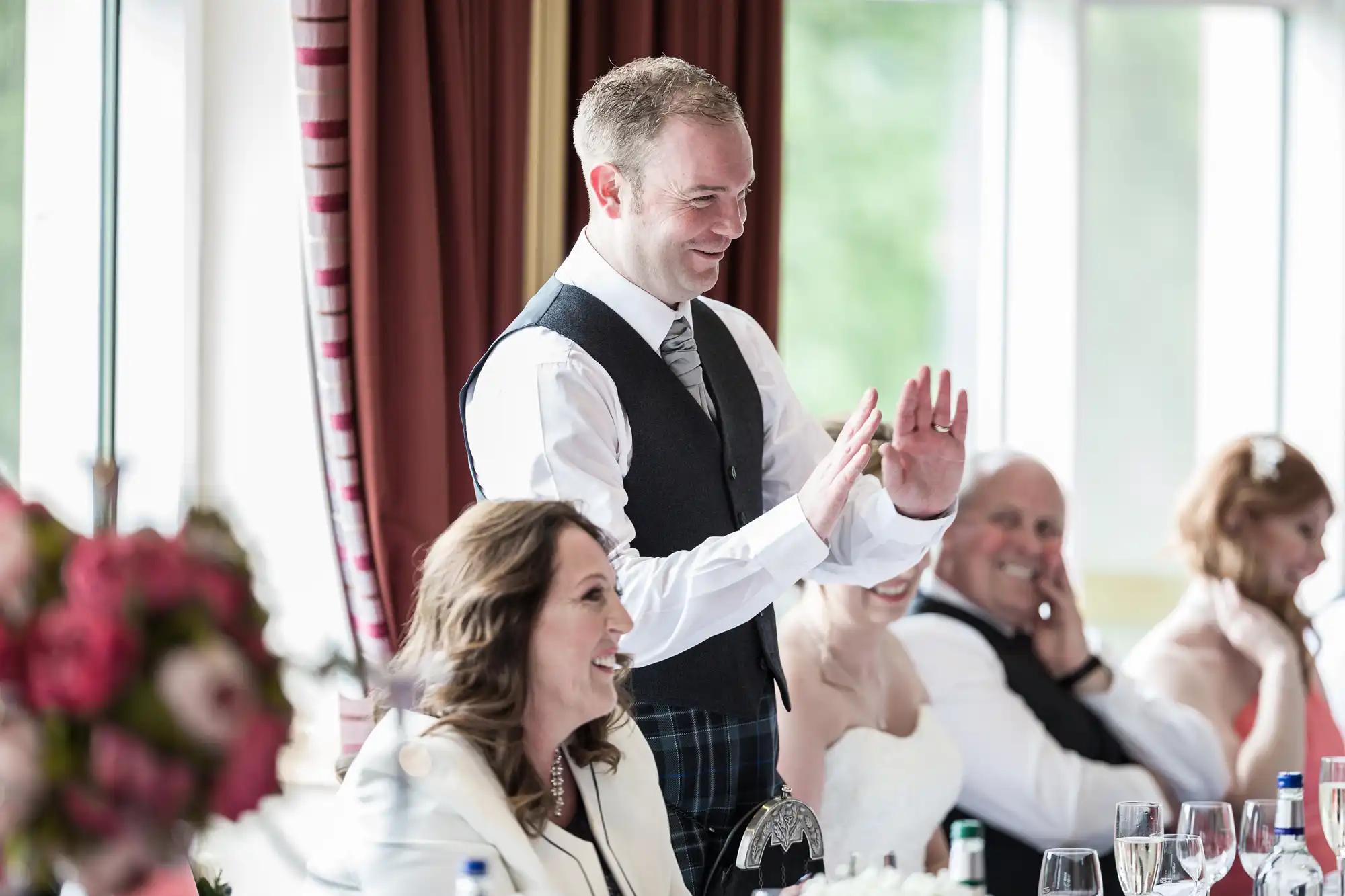 A man in a waistcoat and tie stands smiling, gesturing with his hands, while guests seated at a table, including a woman in white and a bride in a wedding dress, laugh. The setting is bright with large windows and red curtains. Flowers and glasses are visible.
