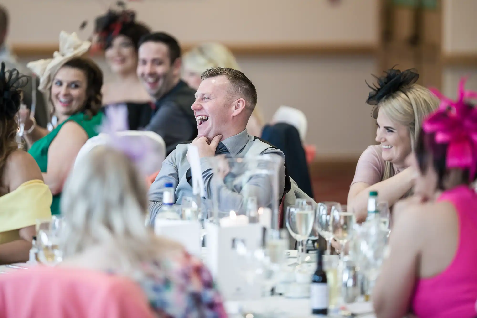 A group of well-dressed guests sit around a table at a formal event, laughing and smiling. The table is set with glasses, bottles, and decorative candles. Many attendees wear colourful outfits and elegant hats or fascinators, creating a cheerful, celebratory atmosphere.