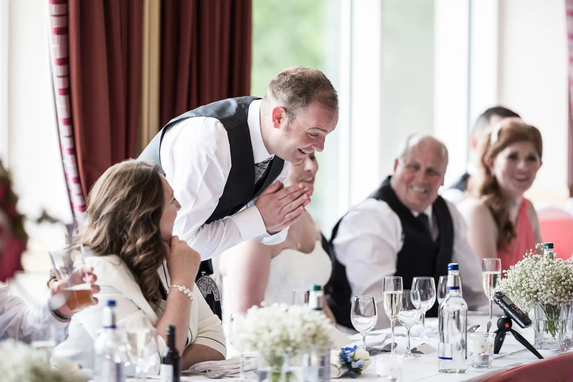A man in a white shirt and black waistcoat stands and smiles with hands clasped, as people sit beside him at a decorated table with drinks, glasses, and flowers. The group appears joyful, likely at a wedding reception with red curtains in the background.