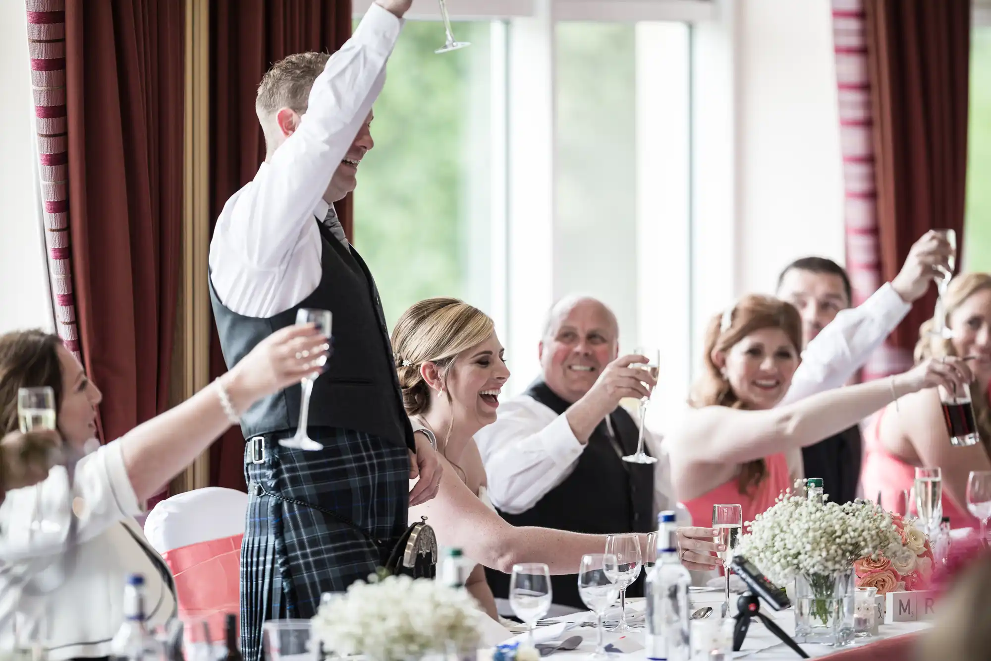 A group of people at a formal event, possibly a wedding, sit at a decorated table raising glasses in a celebratory toast. One man in a kilt stands and leads the toast. The group smiles, surrounded by floral centrepieces and drinks.