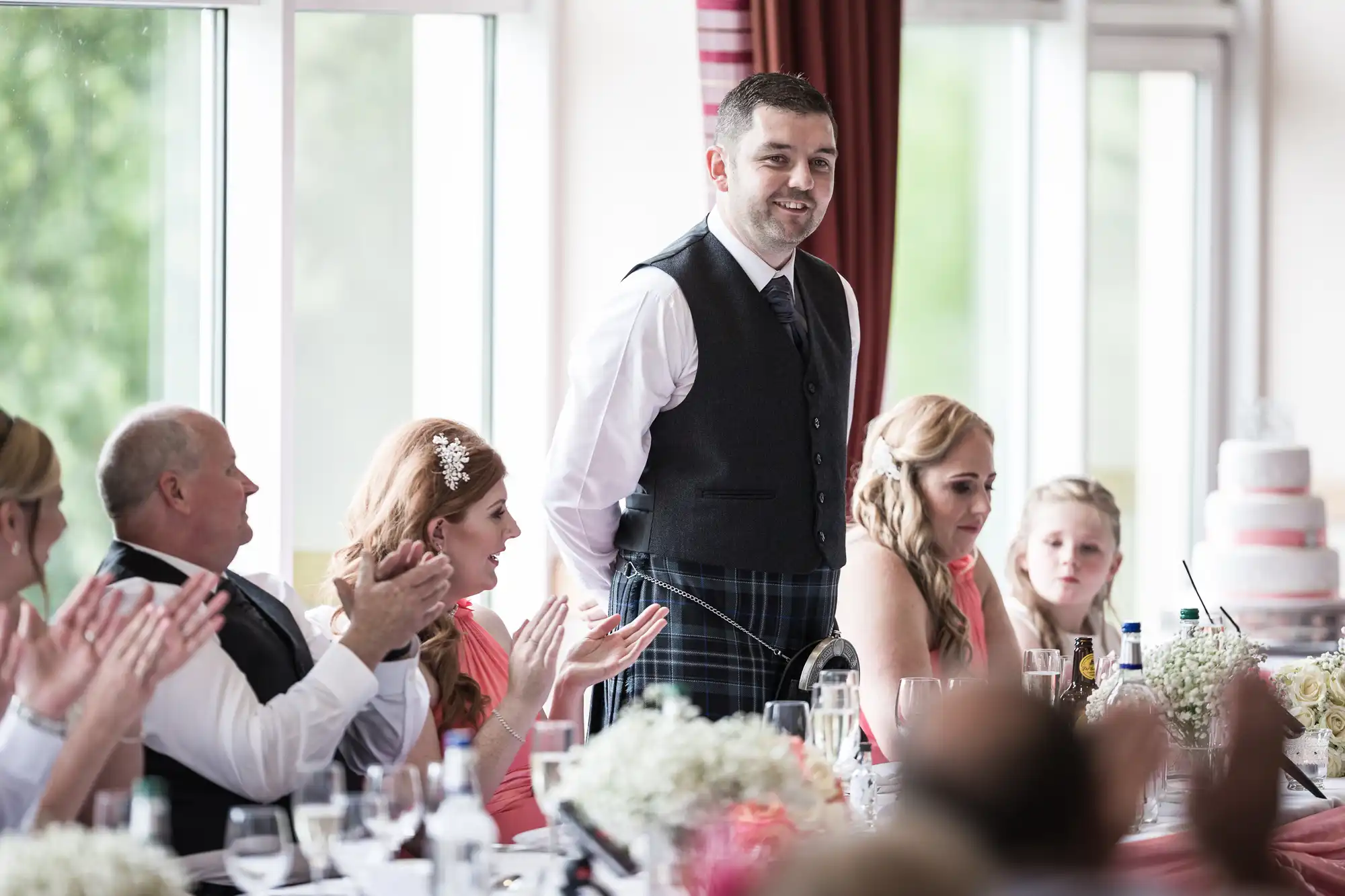 A man in a waistcoat and kilt stands smiling at a wedding reception table. Guests around him, dressed formally, are seated and applauding. The table is set with flowers and glasses. In the background, large windows and a tiered wedding cake are visible.