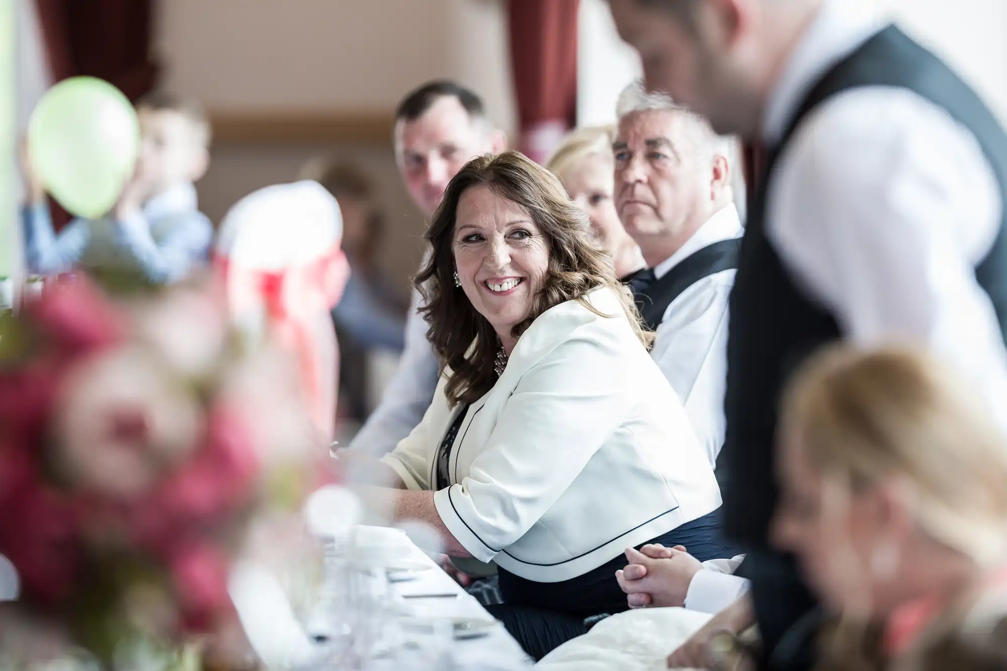 A woman in a white jacket smiles whilst seated at a long table during a formal event. Other guests sit nearby, some blurred in the foreground, all dressed formally. The atmosphere appears celebratory with flowers and soft lighting.