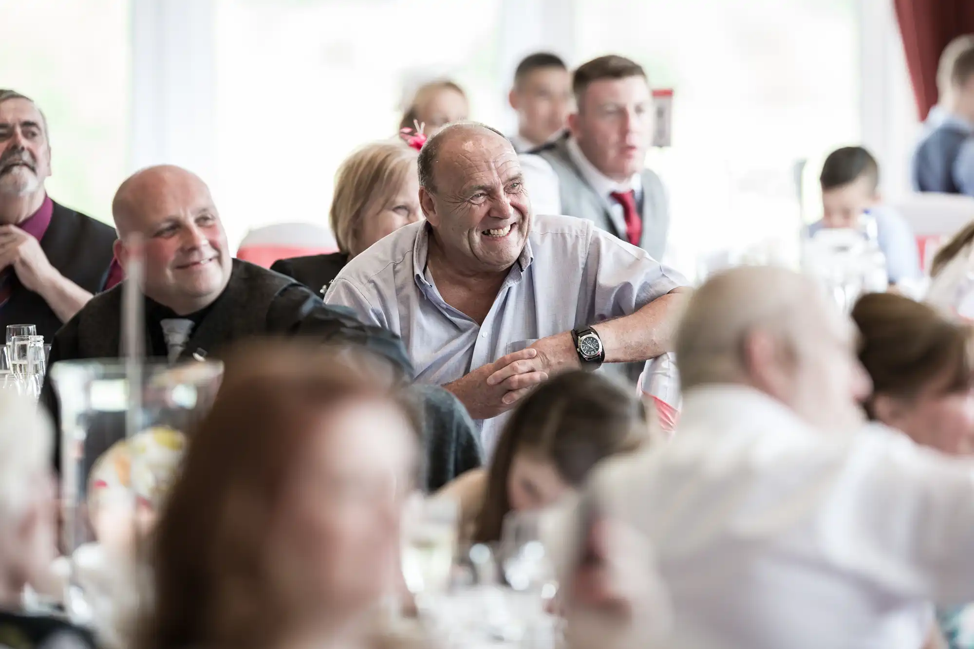 A group of people sit at tables during a formal event. The focus is on a smiling, balding man in a light shirt at the centre. Others around him are blurred, suggesting a lively atmosphere, with some dressed in suits and ties. The background is softly lit.