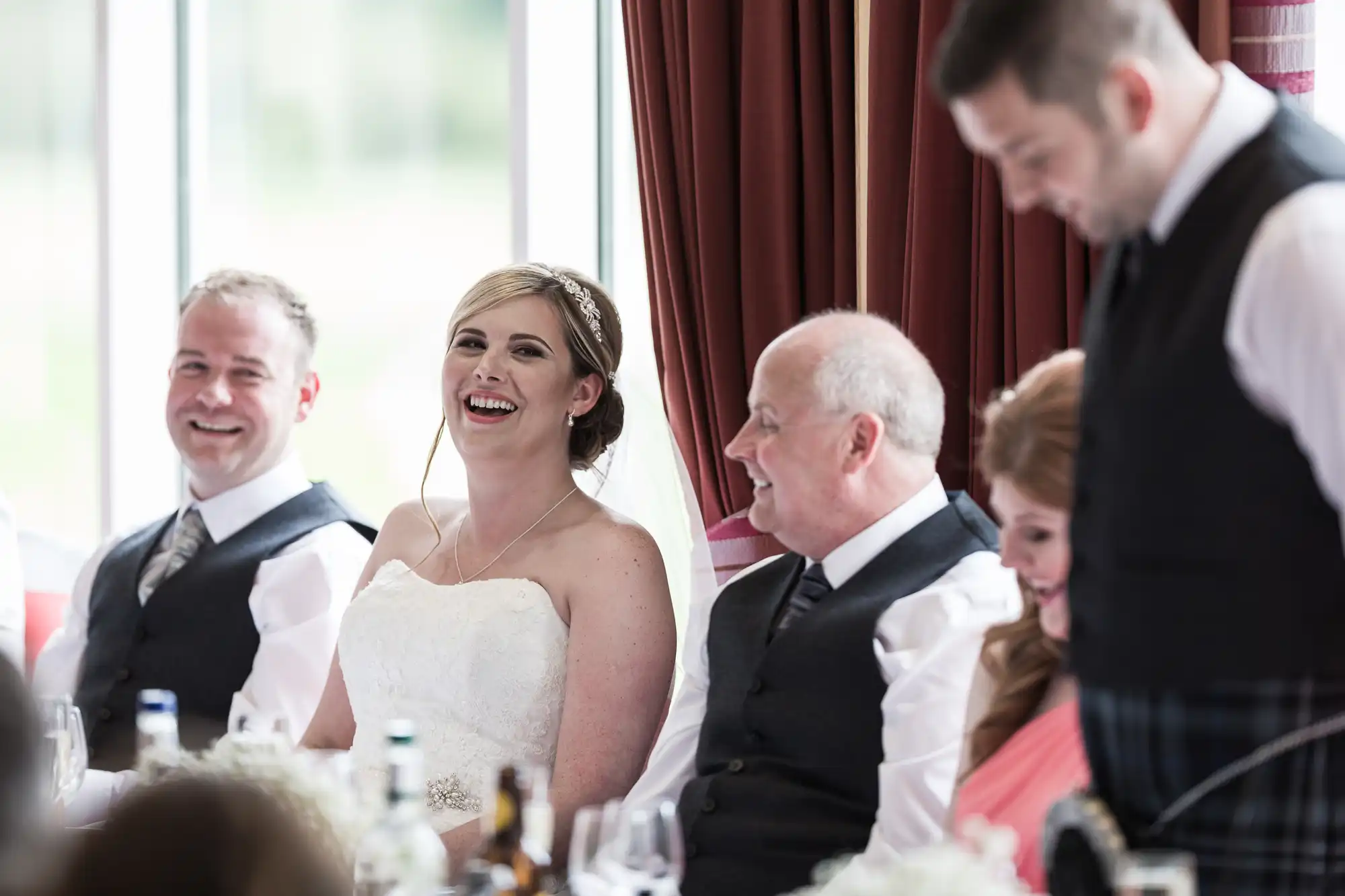 A bride in a strapless white dress and tiara smiles and laughs at a table with three men and a woman in formal attire. They are seated indoors near large windows with maroon curtains, appearing to enjoy a wedding celebration.