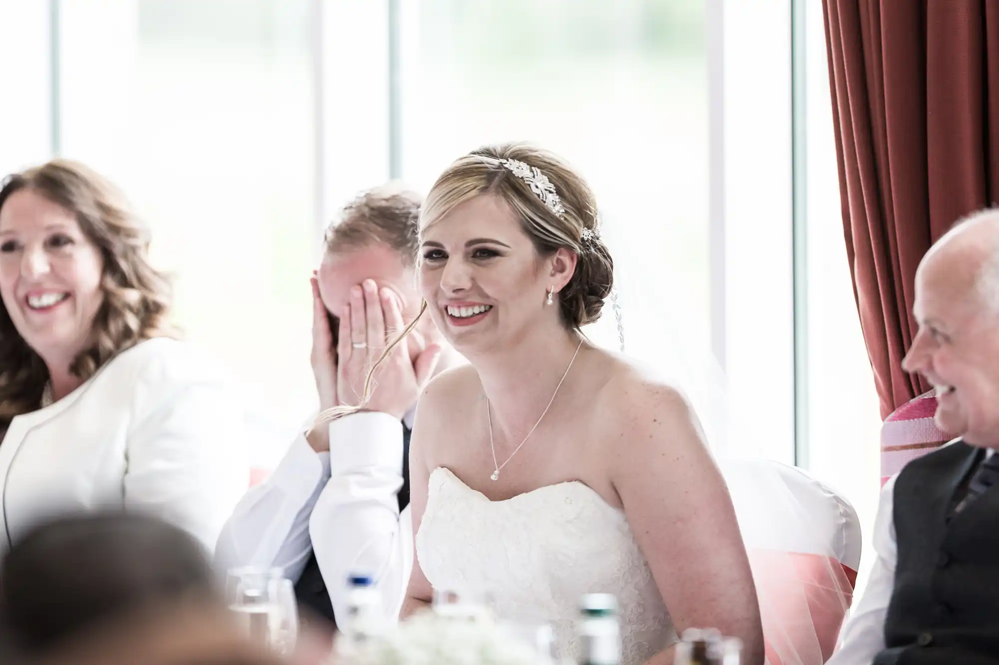 A bride in a white dress smiles at a table, surrounded by wedding guests. A man beside her covers his face with his hand, whilst two other people, also smiling, sit nearby. The background shows large windows with red curtains.