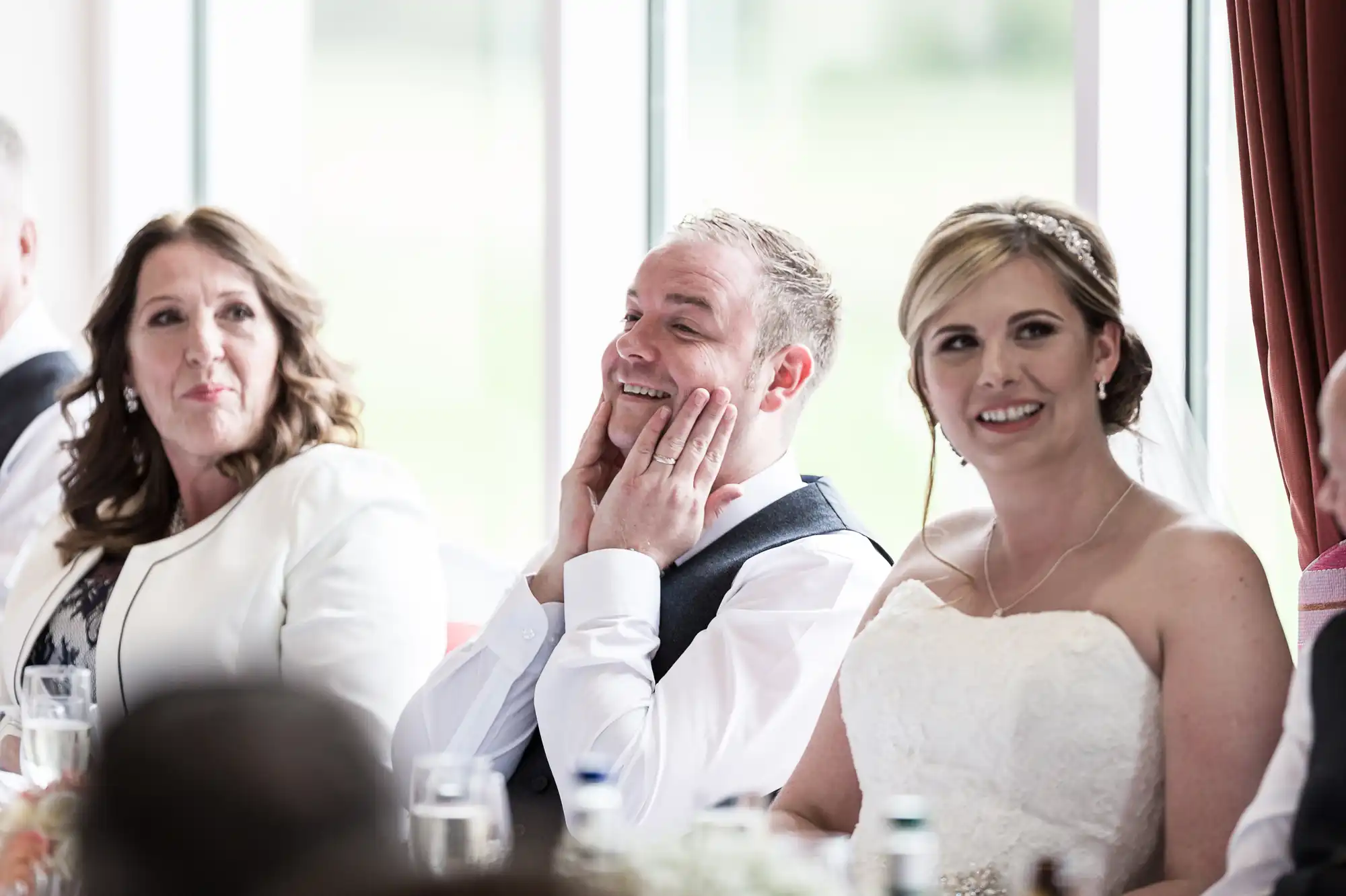 Three people sit at a wedding reception table. The groom, wearing a waistcoat, smiles and holds his face in his hands. The bride in a strapless white dress and tiara sits beside him, smiling. Another woman in white sits to their left, also smiling.
