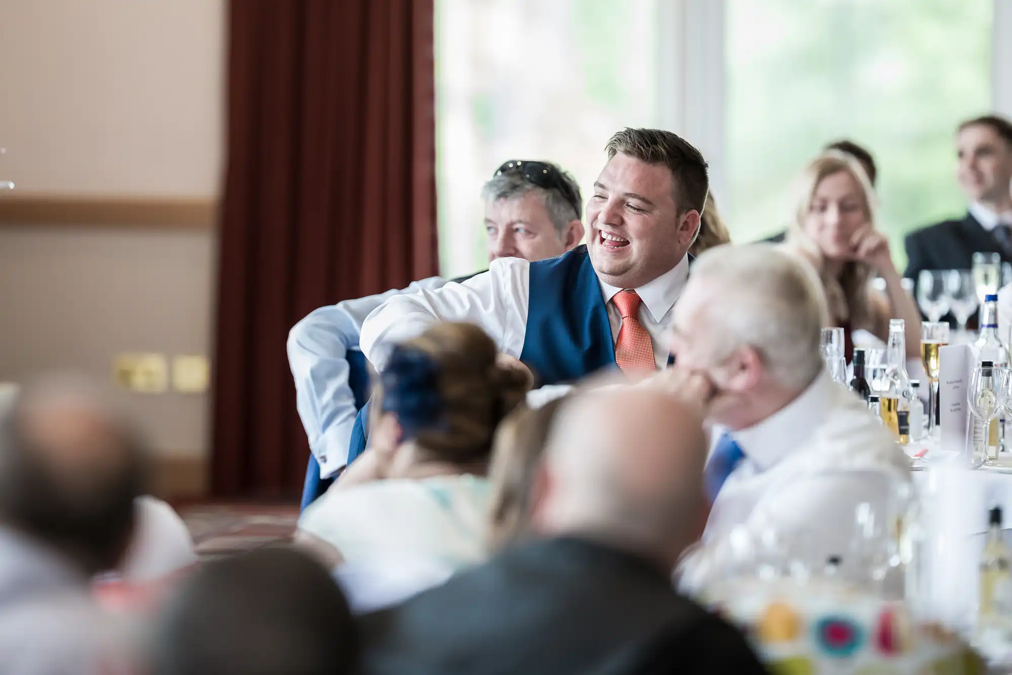 A man in a blue waistcoat and orange tie laughs while seated at a table among other people at an indoor event. Several people are watching him, and bottles and glasses are visible on the tables. The background is softly blurred with natural light coming in.