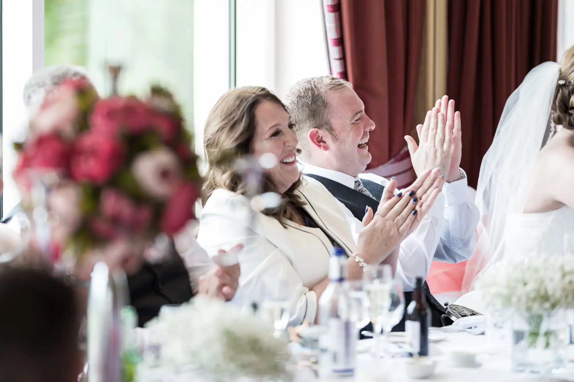 A man and woman dressed formally sit at a table, smiling and applauding, likely at a wedding reception. Flowers, bottles, and glasses are on the table. Another person in a white dress and veil, possibly a bride, sits beside them. The mood is joyful and celebratory.