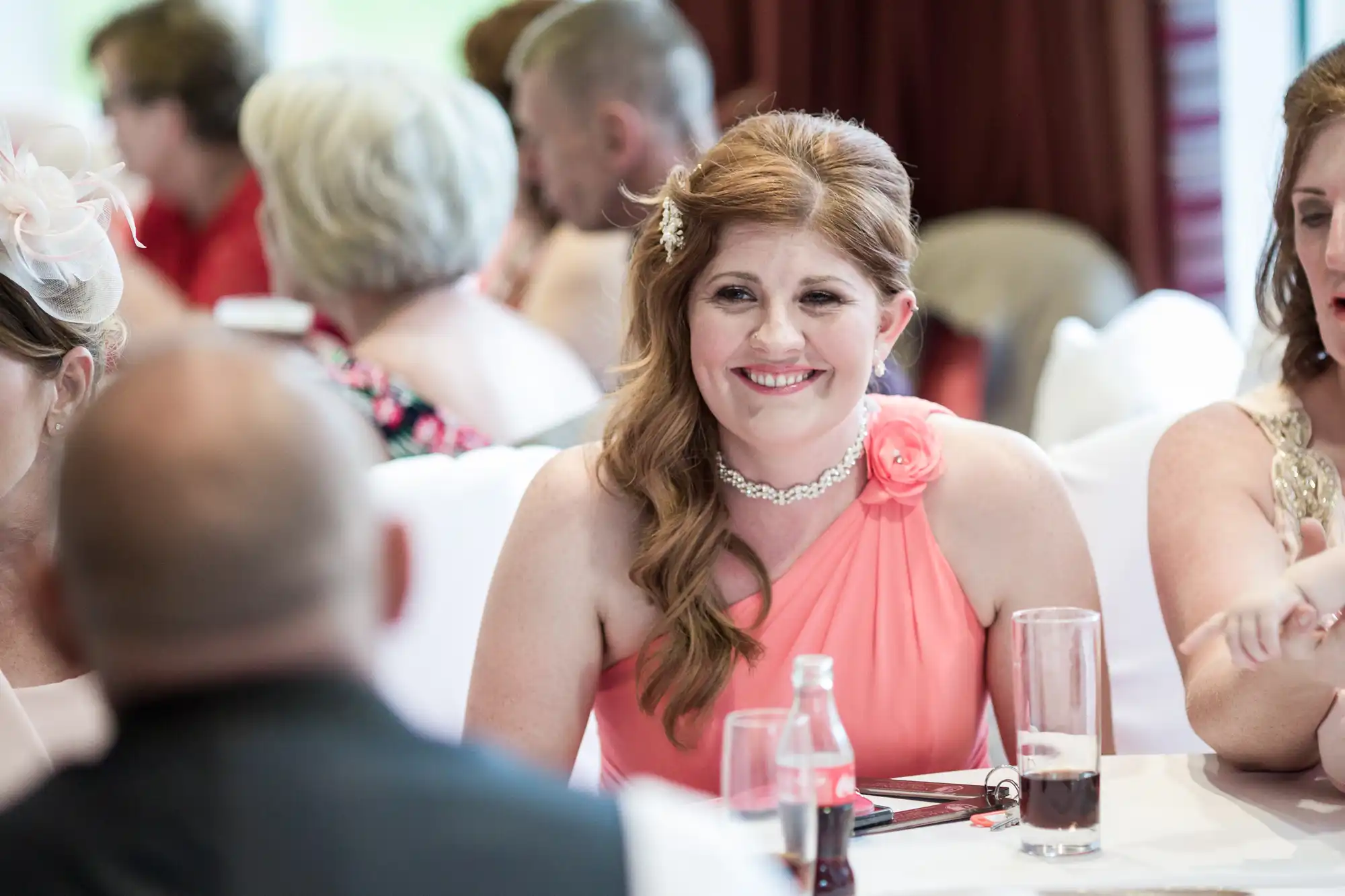 A smiling woman with long red hair, wearing a pink one-shoulder dress and pearl necklace, sits at a table at a formal event. She is surrounded by other people, drinks, and white tablecloths in a brightly lit, indoor setting.
