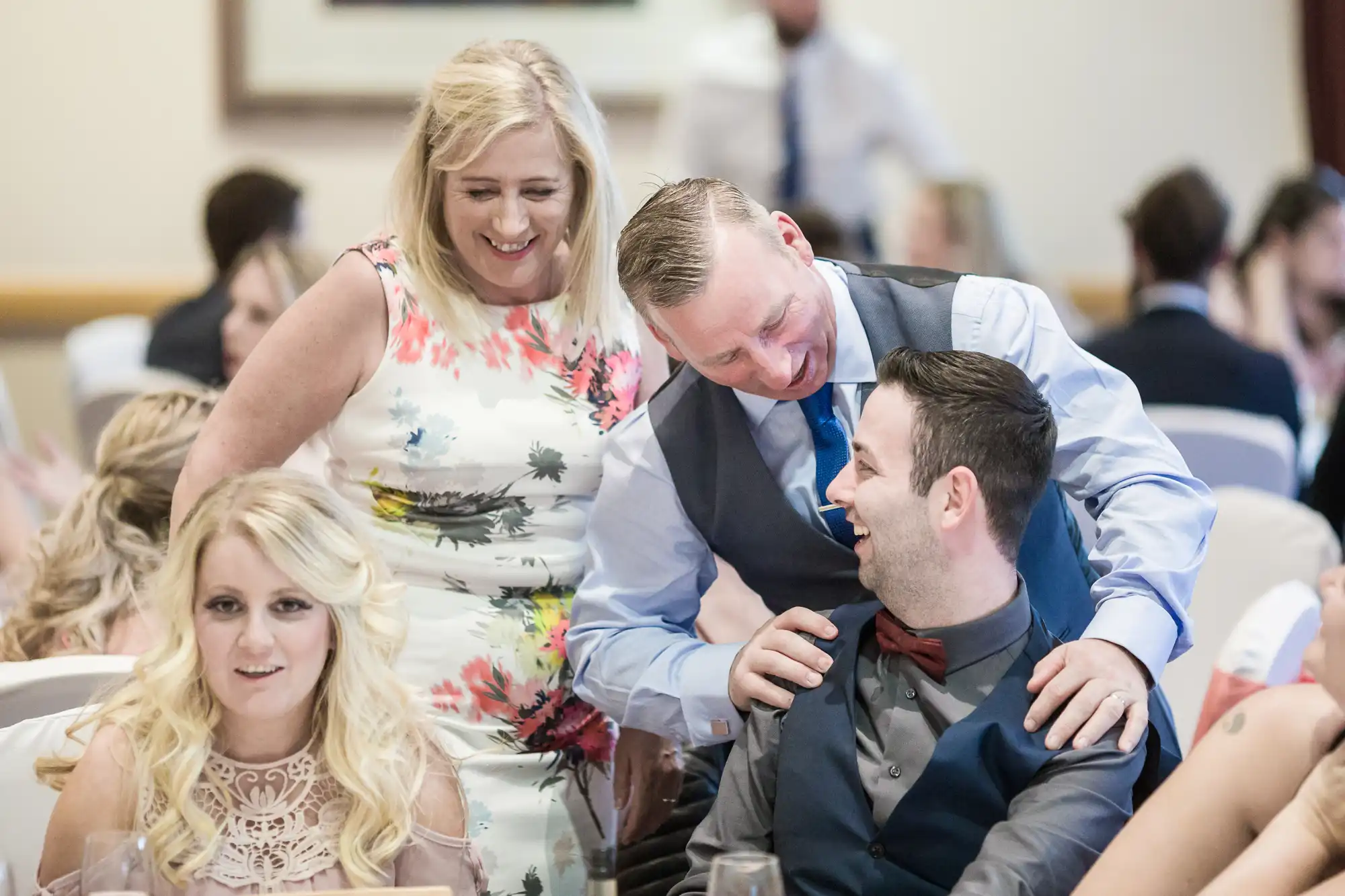 A smiling woman in a floral dress stands by two men at a formal event. One man in a waistcoat and tie leans over and laughs with another seated man in a bow tie. A blonde woman in a light dress sits in the foreground, also smiling. The background is softly blurred.
