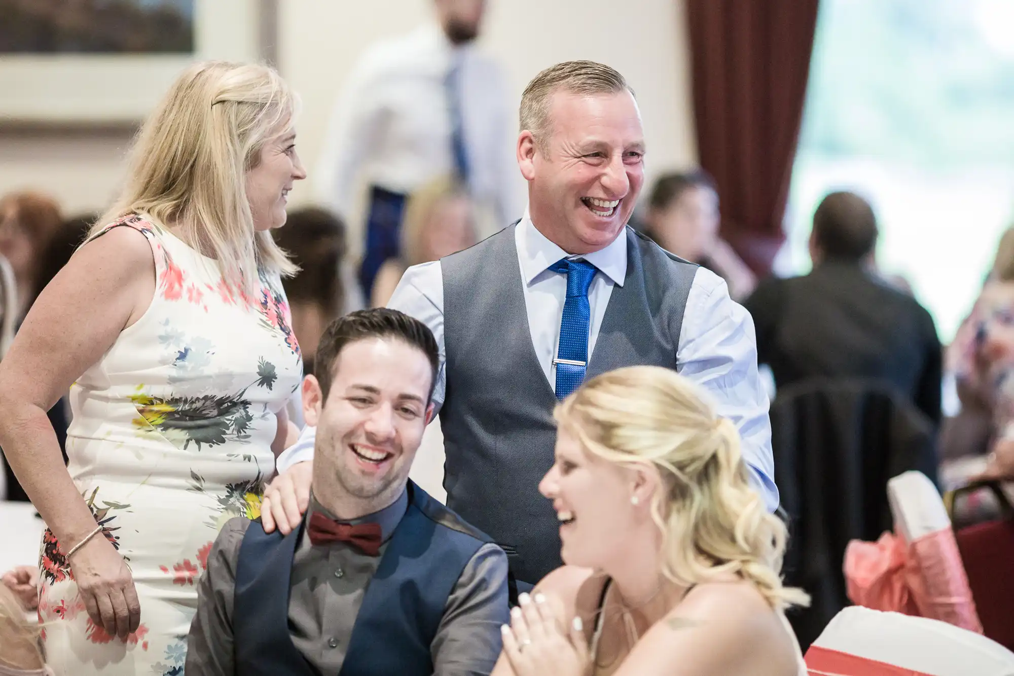 Four people at a lively indoor event. Two are standing, smiling, and talking to two seated guests. All are dressed in formal attire, and the atmosphere appears joyful, with blurred people and tables in the background.