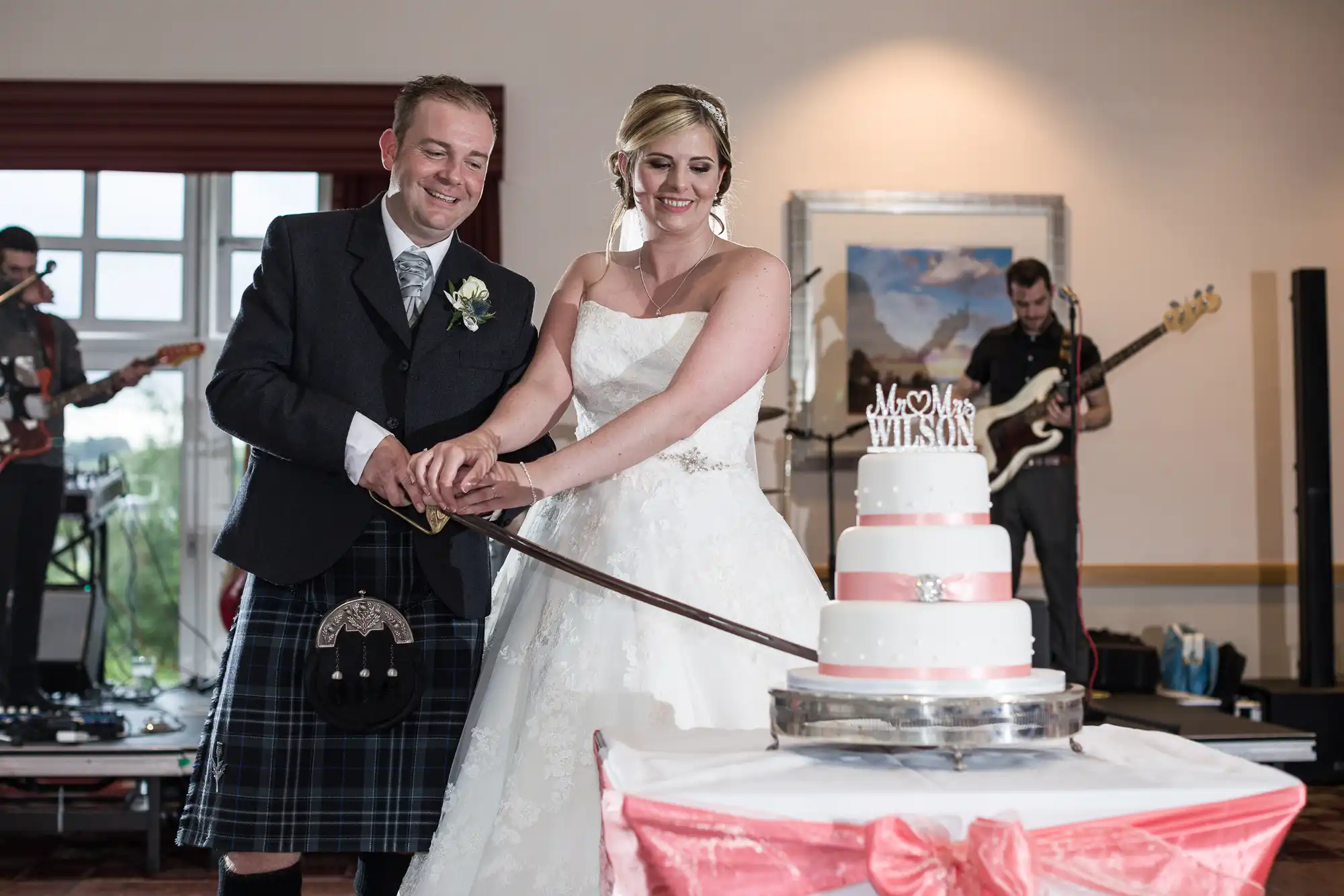 A smiling bride in a white gown and a groom in a kilt cut a white and pink wedding cake with a sword. A band plays in the background. The cake has a “Mr & Mrs Wilson” topper and sits on a table draped with a pink cloth and bow.
