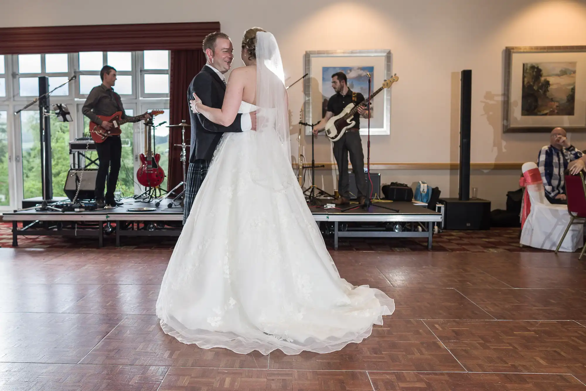 A bride in a white dress and veil dances with a groom in a suit on a wooden floor. A live band with guitars and drums performs in the background. Framed art hangs on the wall, and a seated guest in red and white watches. Large windows let in daylight.